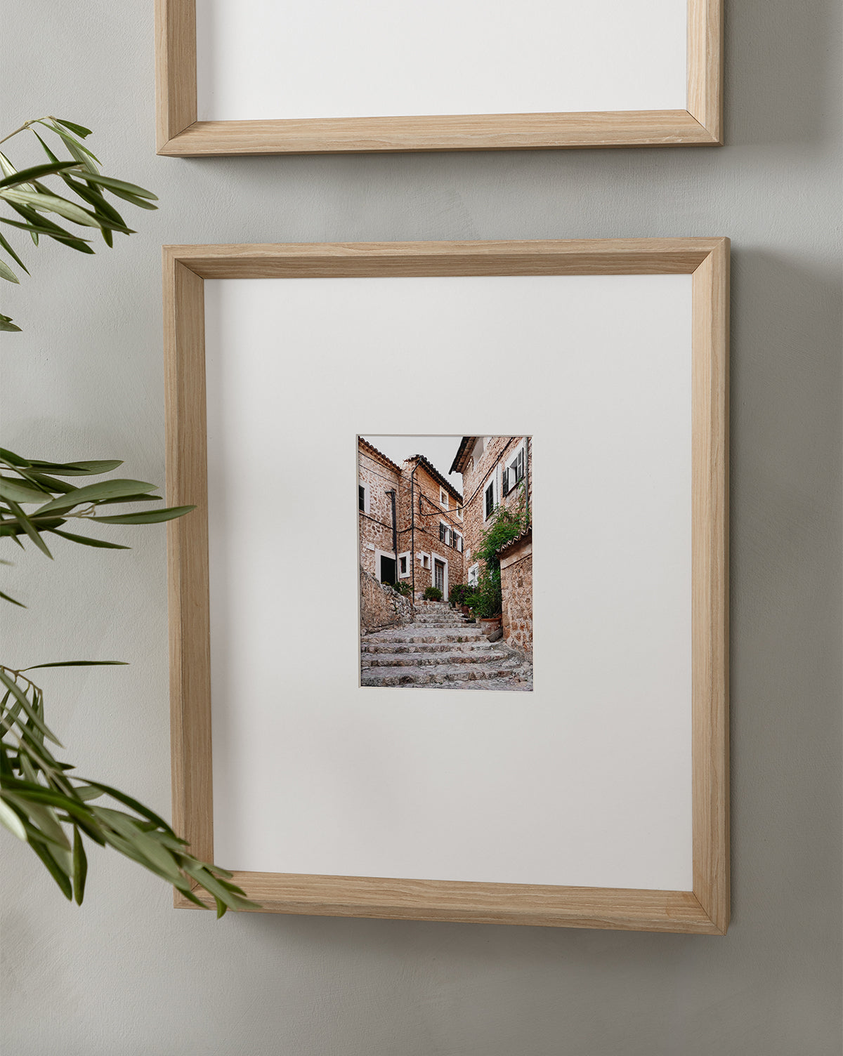 The Fornalutx framed photo highlights expert composition of a rustic, stone-paved alley with old brick buildings and greenery, hung on a light wall, partially overlapped by another frame above and leaves in the foreground.