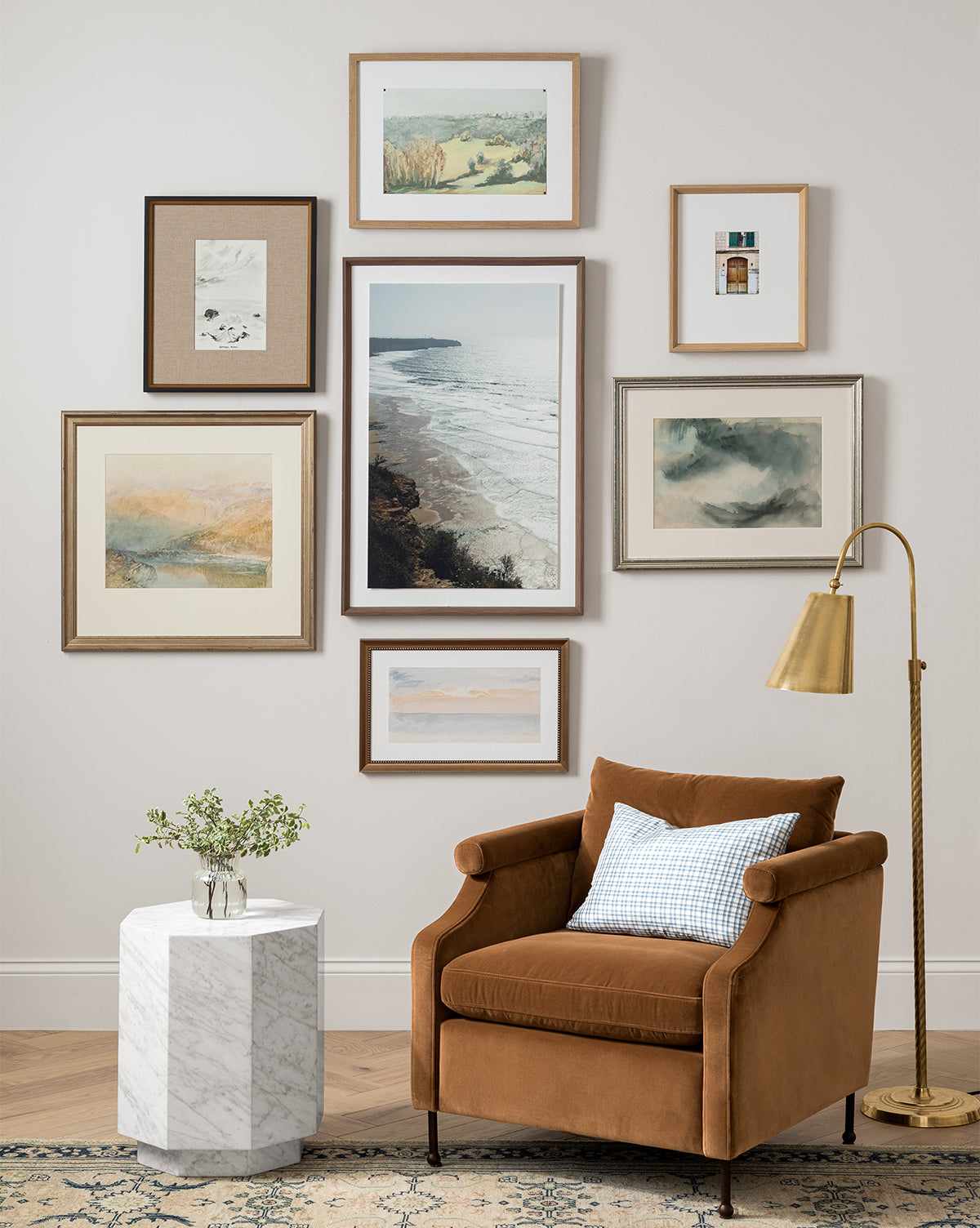 A cozy corner with a rust-colored armchair, patterned pillow, white marble side table with greenery, and gold floor lamp is finished by a gallery wall featuring the Town of Deia giclee print landscape art.