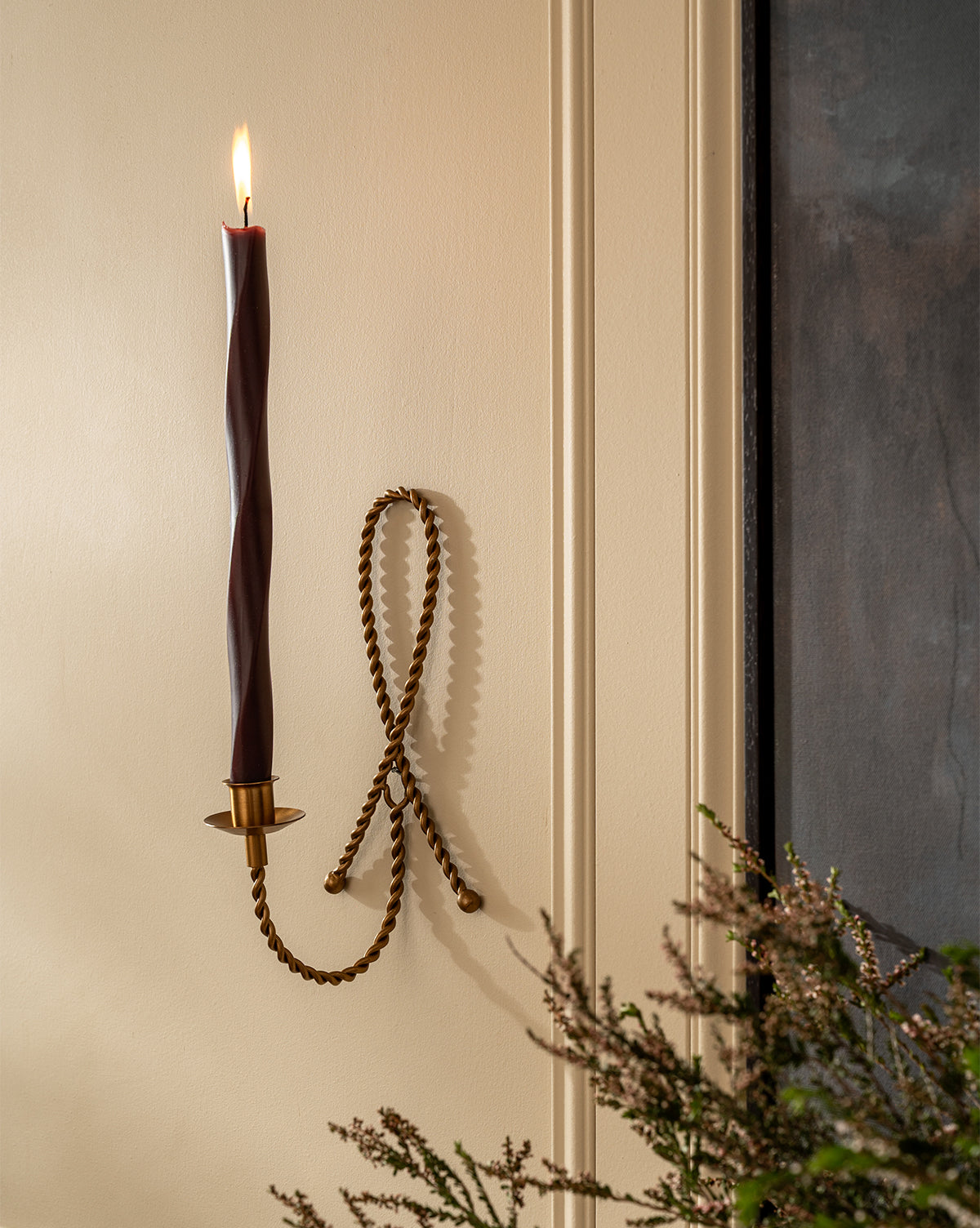 A lit twisted burgundy candle is placed in the Hearst Taper Holder, a sculptural brass piece with a decorative loop, mounted on a cream wall. Greenery appears in the bottom right corner, by McGee & Co.
