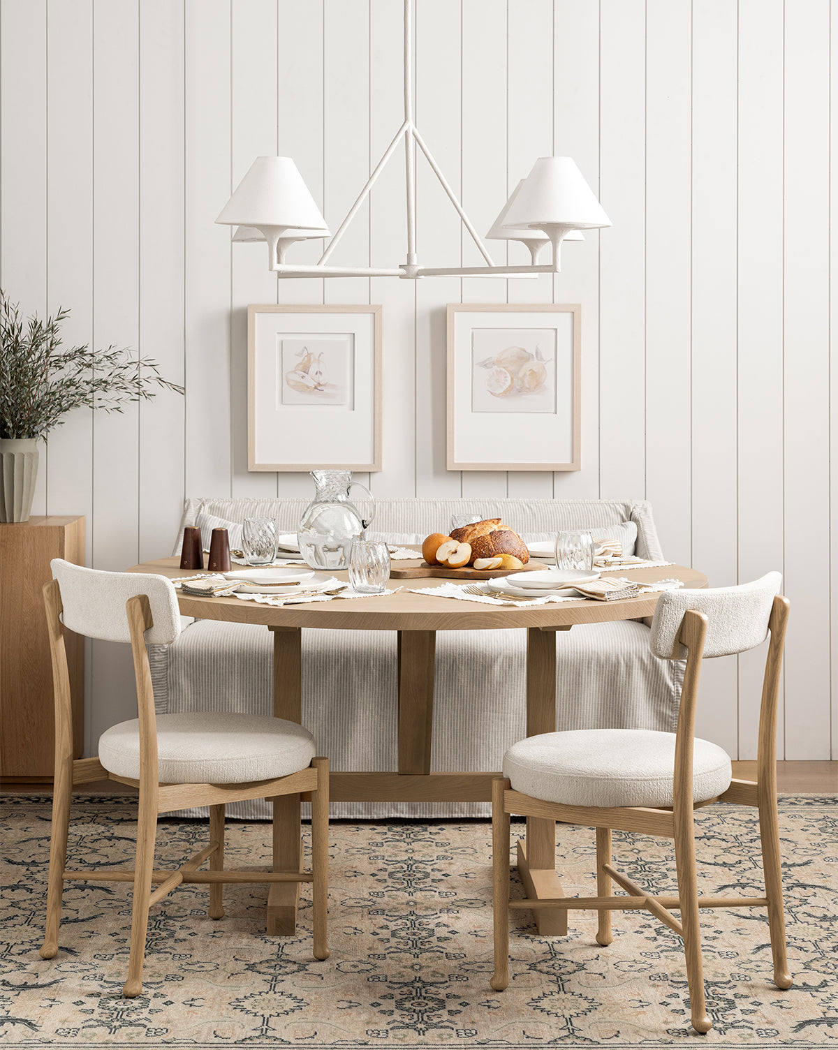 A cozy dining area featuring a Giselle Round Table in kiln-dried white oak, two cushioned chairs, a patterned rug, a striped bench cover, wall art, and a white three-light chandelier against paneled walls by McGee & Co.