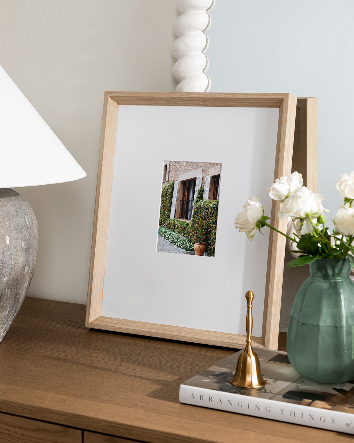 A wooden dresser with a lamp, the La Residencia Deia framed giclee print, a green vase with white roses, a gold bell, and books—all reflected in a scalloped-edge mirror with a natural woodgrain frame.