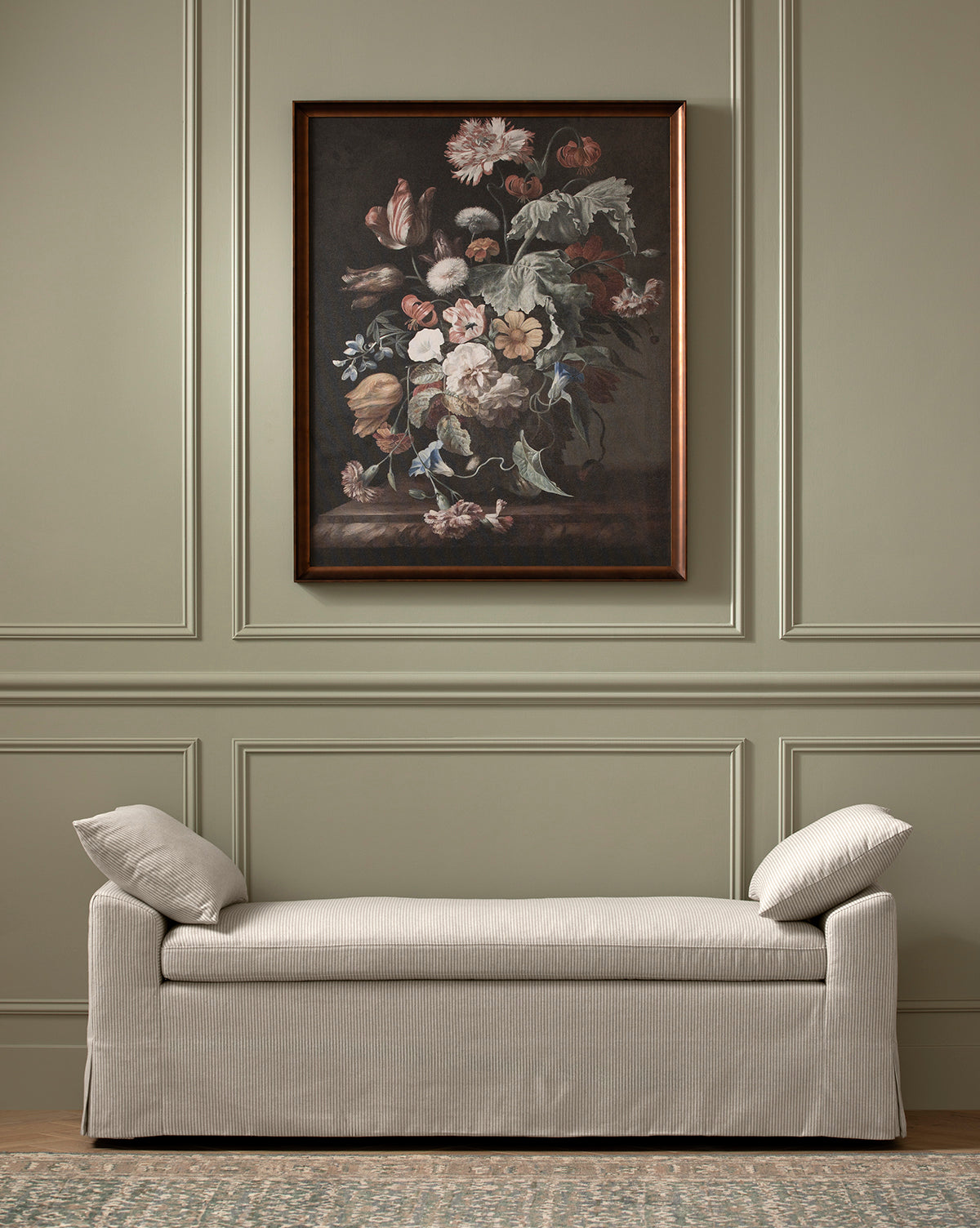 A beige upholstered bench with two cushions sits against a light sage paneled wall. Above it hangs the Botanical Renaissance floral still life in a bronze-finished frame. A patterned rug partially covers the wooden floor.