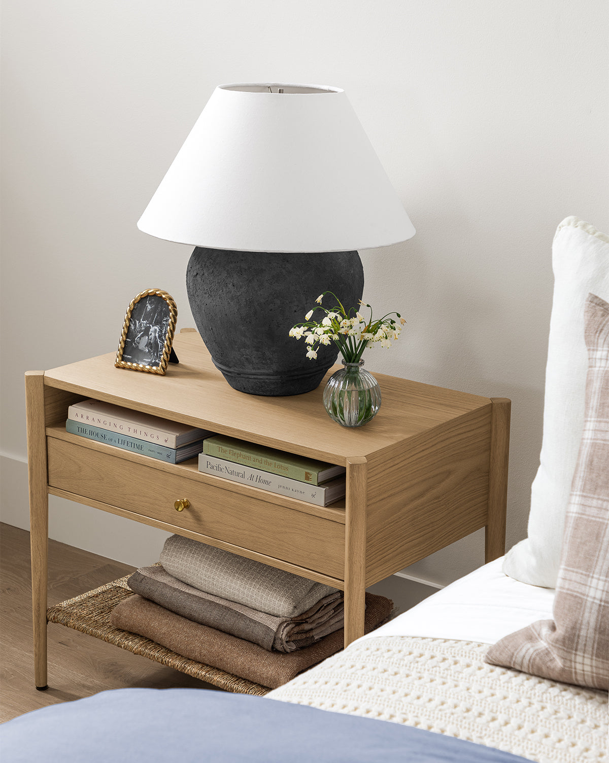 A wooden bedside table holds a black lamp with a white shade, books, a framed photo, and decorative vignettes including flowers in the Verlice Glass Round Vase. Folded blankets rest on the lower shelf beside the bed’s plaid pillow.