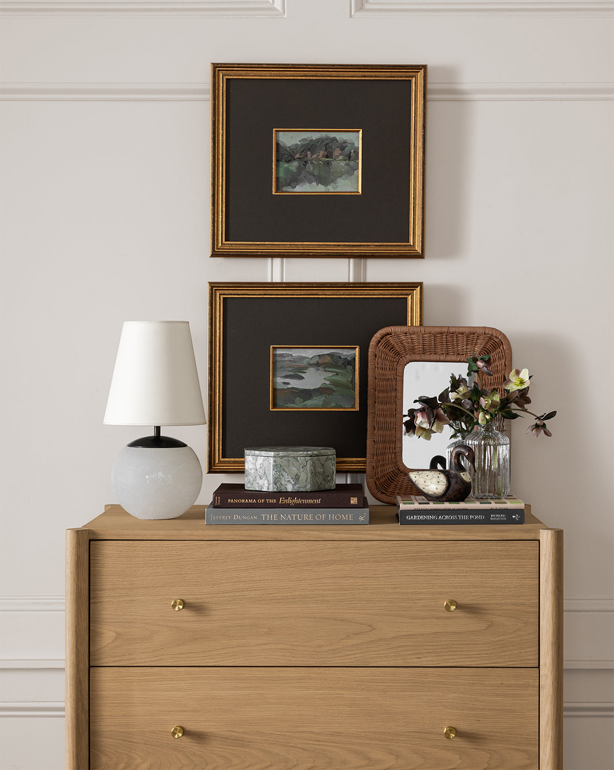 A wooden dresser with gold knobs displays a white lamp, books, green box, wicker-framed mirror, and a flower vase. Above it hang two framed paintings, one of which is Sage Shores II in an antique gold frame.