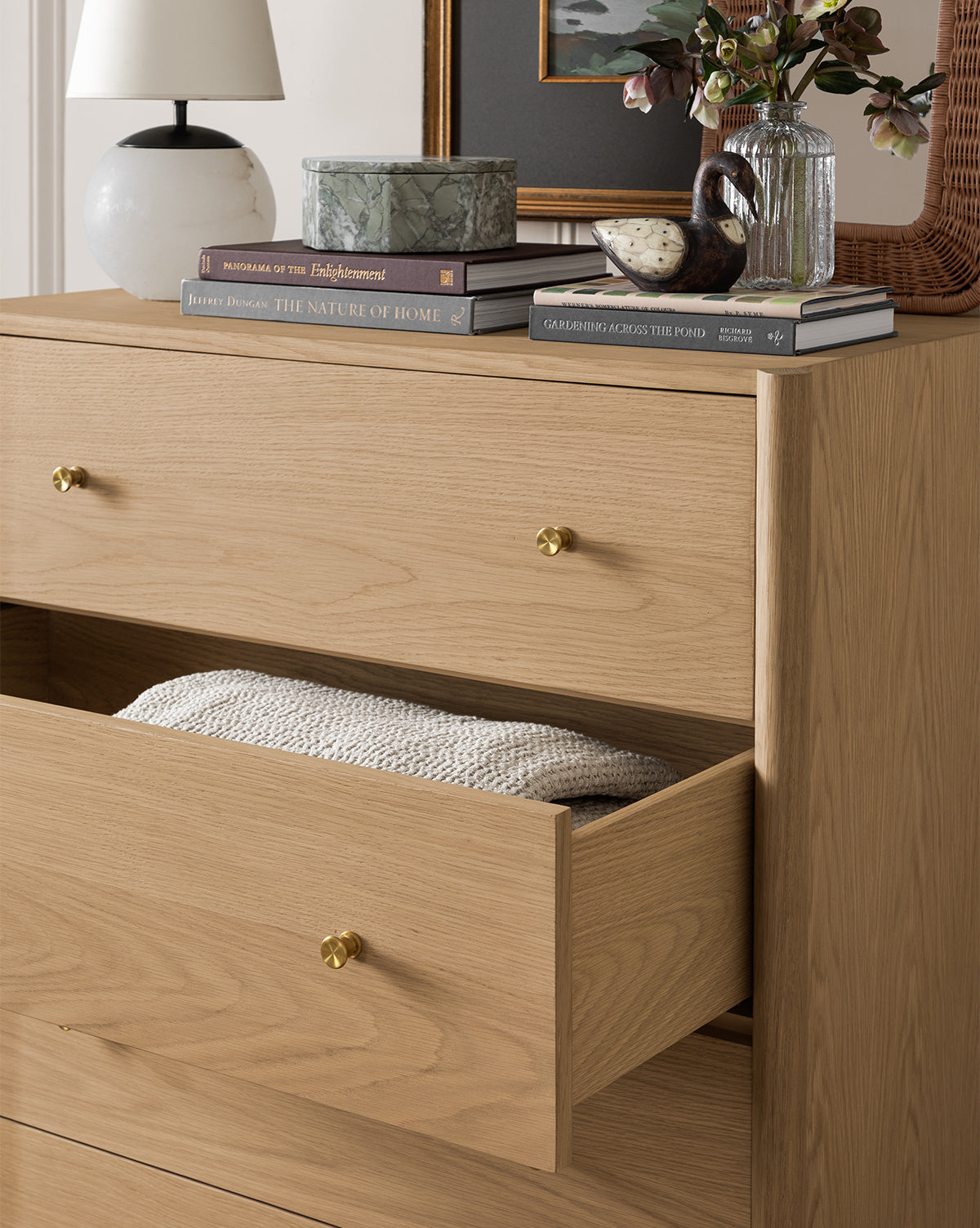 The Dana Tallboy Dresser features white oak and brass hardware, with a partially open drawer revealing a folded knit blanket. On top, find stacked books, a lamp, flowers in a vase, a ceramic bird, and a framed picture on the wall above by McGee & Co.