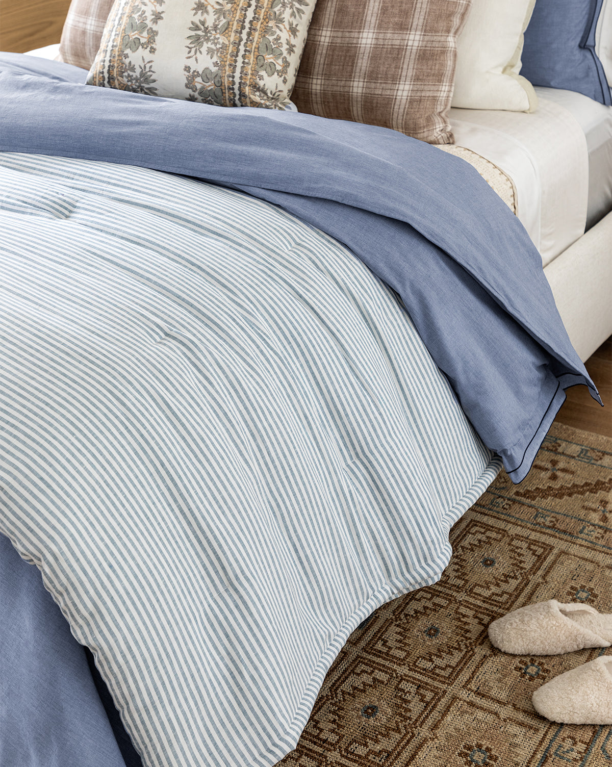 A neatly made bed with blue and white striped linen, the Rosemary Quilt, decorative plaid and floral pillows, and beige slippers on a patterned rug beside the bed.