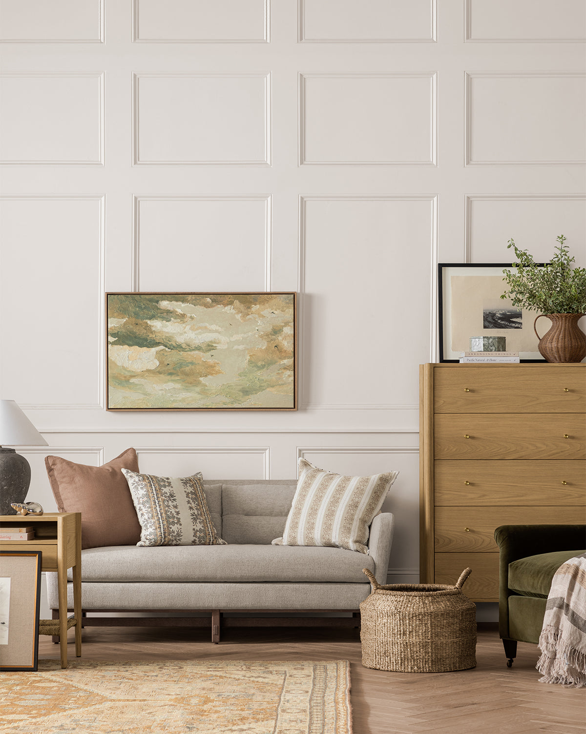 A cozy living room with a light gray sofa, patterned pillows, wooden side table, basket, tall dresser with decor, and the Swallows at Dusk landscape canvas in a white oak frame on neutral paneled walls by McGee & Co.