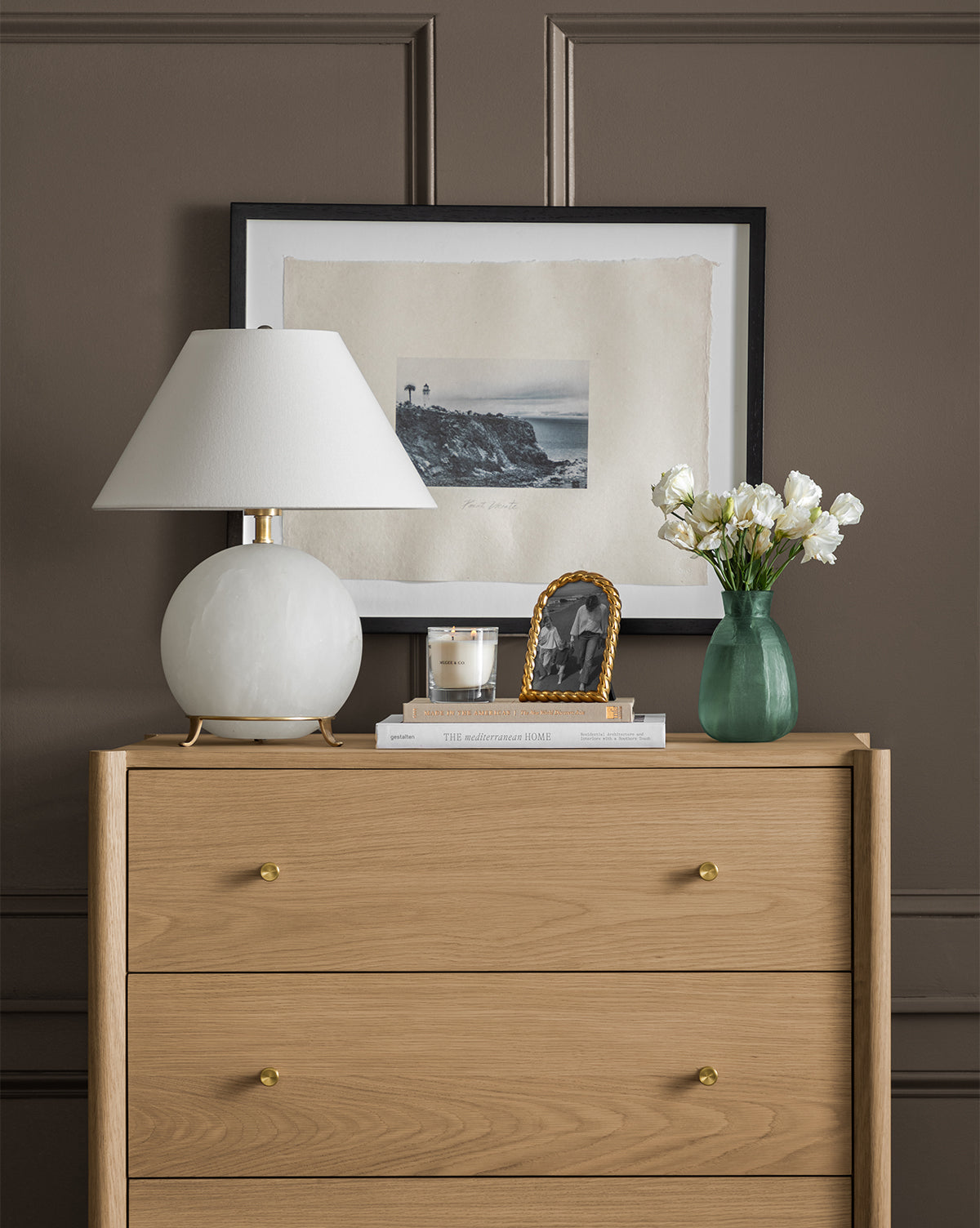 A modern wooden dresser with gold knobs displays the Price Table Lamp with a linen shade, a candle, stacked books, a small framed photo, and a green vase of white flowers. A landscape artwork hangs on the brown wall behind, by McGee & Co.