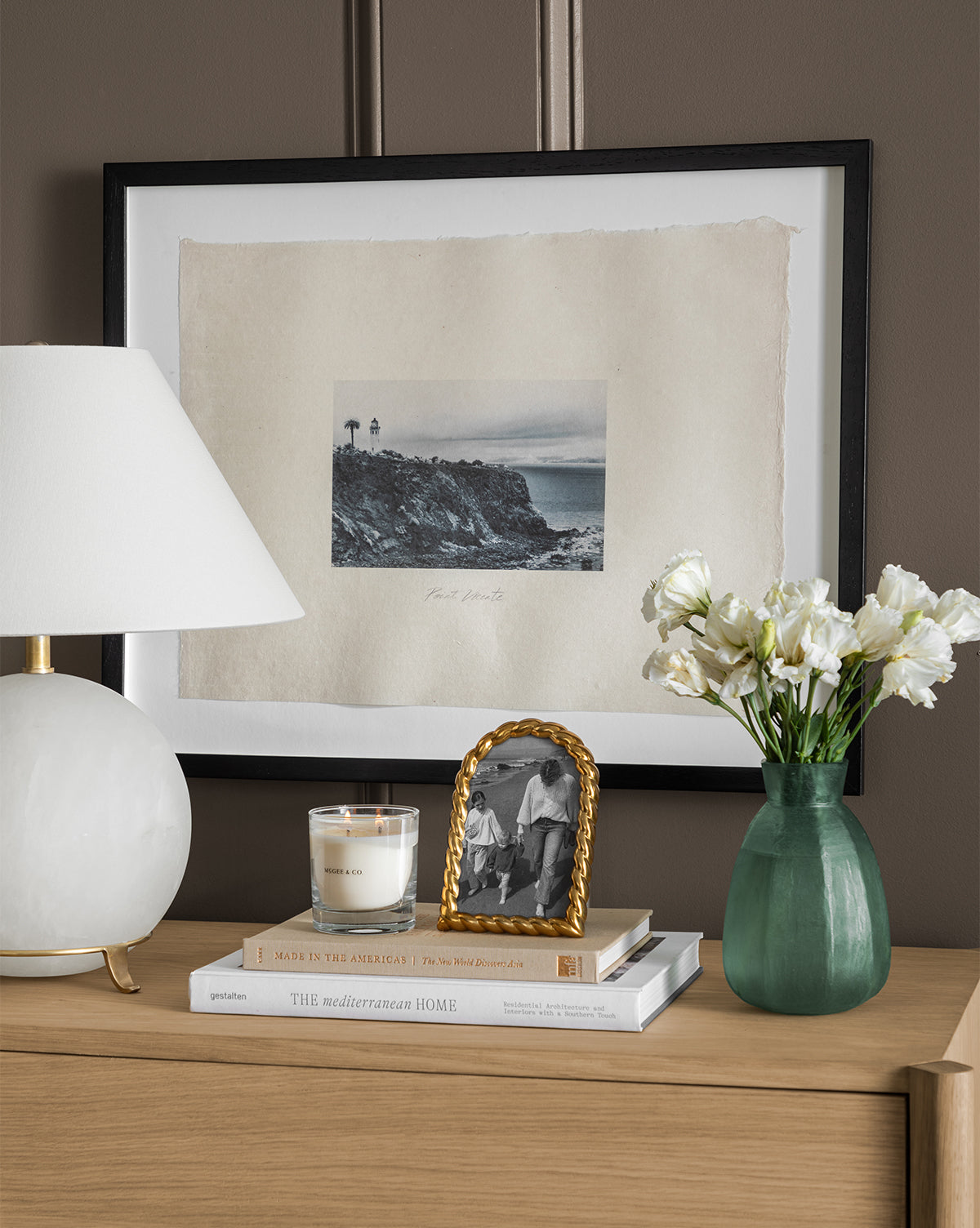 A wooden dresser displays a white lamp, candle, books, photo in gold frame, and green vase with white flowers; behind it hangs Point Vicente vintage wall art on handmade paper against a brown wall.