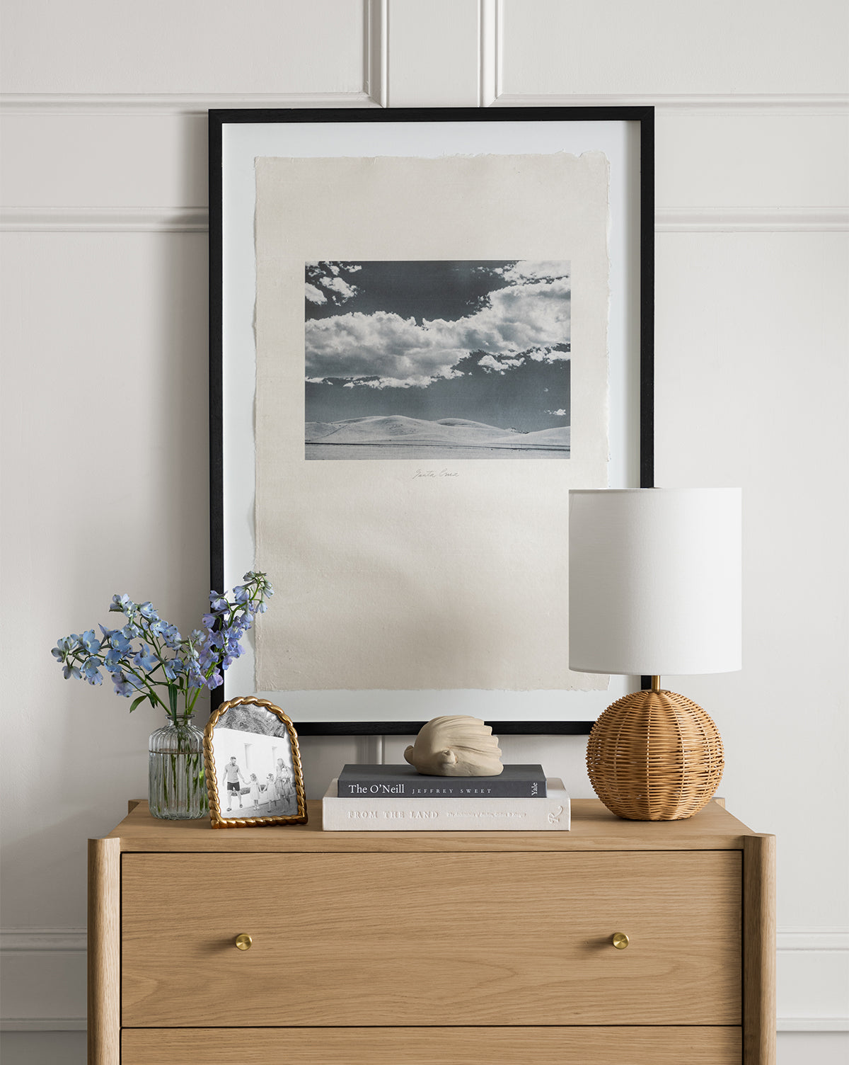 A wooden dresser with gold knobs displays a Mari Table Lamp with a white linen shade and rattan base, blue flowers in a vase, a framed photo, sculpture, and two books. Above is a framed black-and-white landscape photo by McGee & Co.