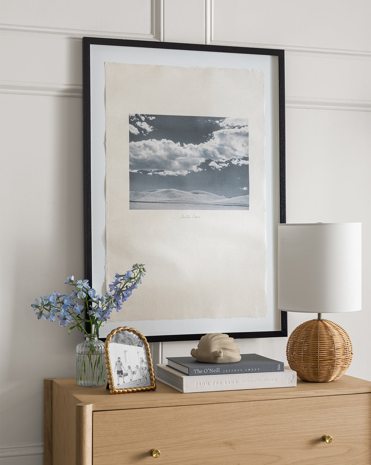 The Santa Cruz dresser features a framed photo, blue flower vase, stacked books, a decorative sculpture, and a rattan lamp. Above it hangs vintage wall art—a large black-and-white sky photo in a classic black frame.