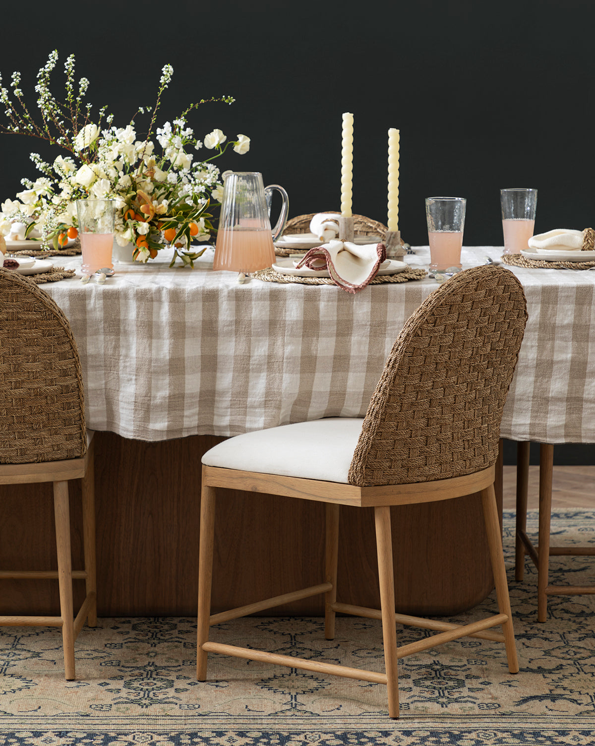A dining table with a beige checkered tablecloth is set with Molly Dining Chairs, a floral centerpiece, tall candles, pink drinks in glasses, a glass pitcher, and napkins for an inviting and elegant look by McGee & Co.