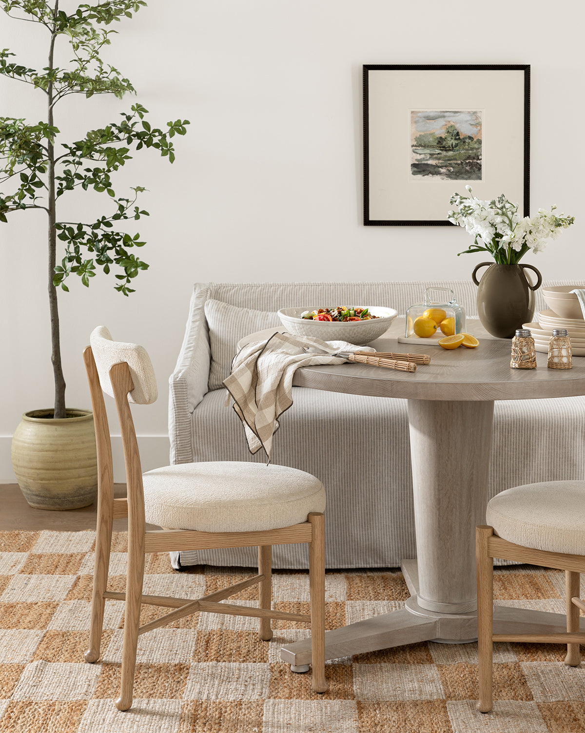 A round wooden dining table with two cushioned chairs, a salad bowl, lemons, and a vase of flowers sits in front of a striped sofa. The cozy dining area is brightened by the Forest Meadow print in a black wood frame on the wall.