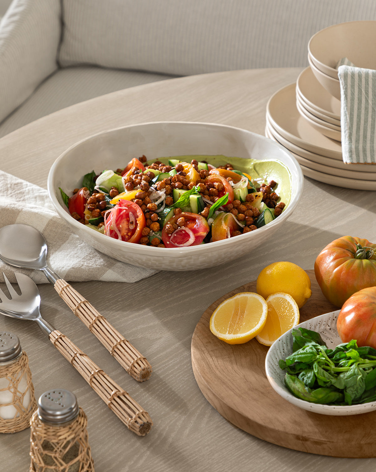 A vibrant salad featuring tomatoes, cucumbers, chickpeas, and greens is served in the Dion Serving Bowl, a ceramic bowl with a white glaze and woven basket design, placed beside lemon halves, basil leaves, plates, utensils, and napkins by McGee & Co.