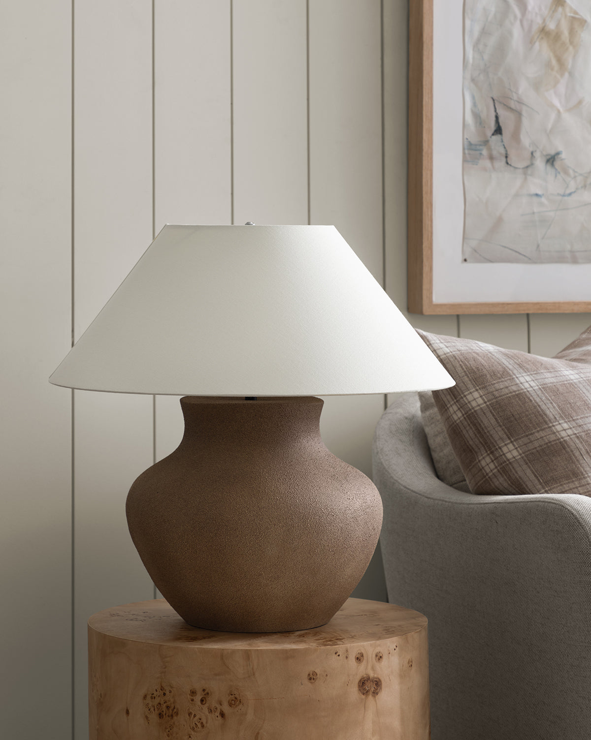 The Parma Table Lamp, featuring a textured ceramic base and ivory linen shade, sits on a wooden side table beside a light gray couch and framed abstract art on a paneled wall, by McGee & Co.