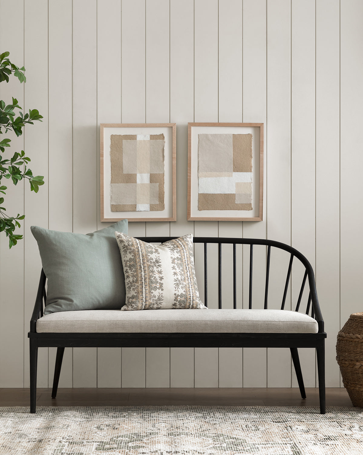 A minimalist living space with a black wooden bench and neutral cushions, Collage 2 handmade paper art in a maple frame on a white shiplap wall, plus a woven basket and a leafy green plant in the corner.