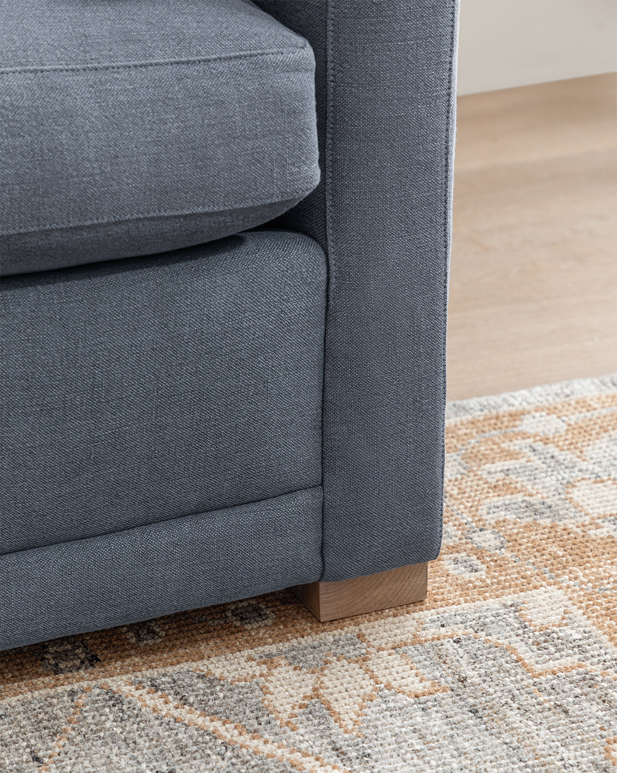 Close-up of the bottom corner of the Peterson Classic Upholstered Sofa (75 - 100) with gray fabric and wooden legs, placed beside a beige and light brown patterned rug on a light wood floor, by McGee & Co.