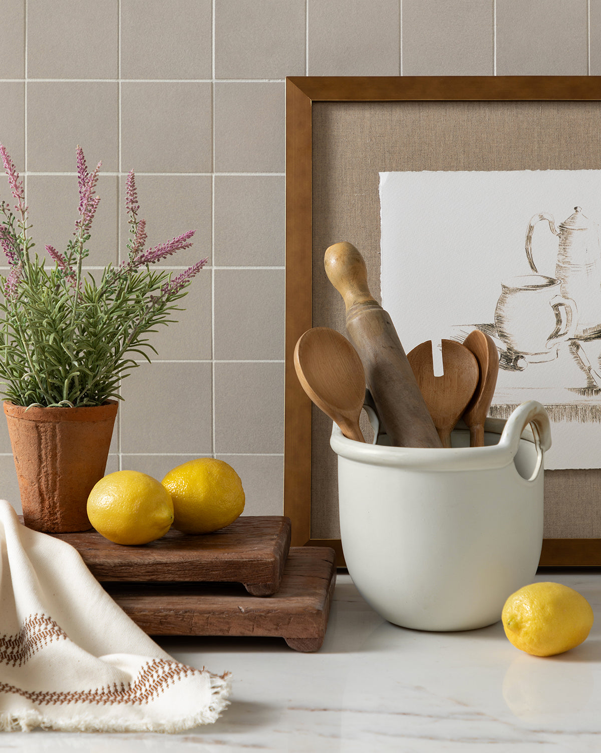 A kitchen counter displays lemons, wooden utensils in a white ceramic crock, a Potted Faux Lavender Topiary, stacked wooden boards, a beige towel, and framed art against a tiled wall.