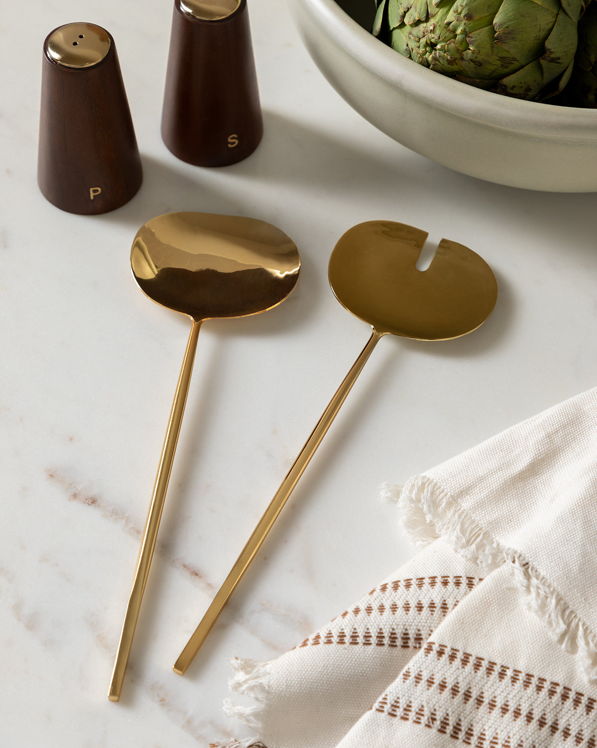 A Woven Stripe Tea Towel is displayed on a marble surface with gold salad servers, wooden salt and pepper shakers, and artichokes in the background, perfect for enhancing your kitchen textiles collection by McGee & Co.