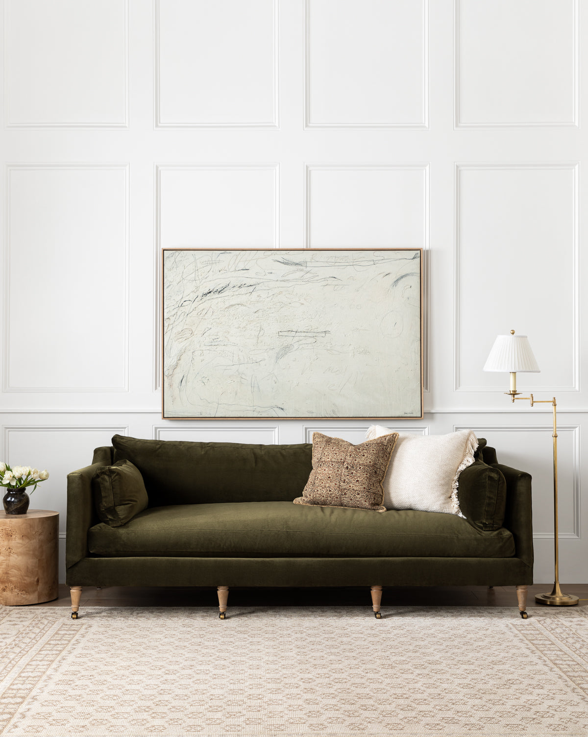 A living room featuring the Rowe Fine Furniture Haverford Upholstered Sofa 90 in olive green velvet with three assorted pillows, a round wooden side table with flowers, a floor lamp, and an abstract painting above a light patterned rug, blending seamlessly with McGee & Co.