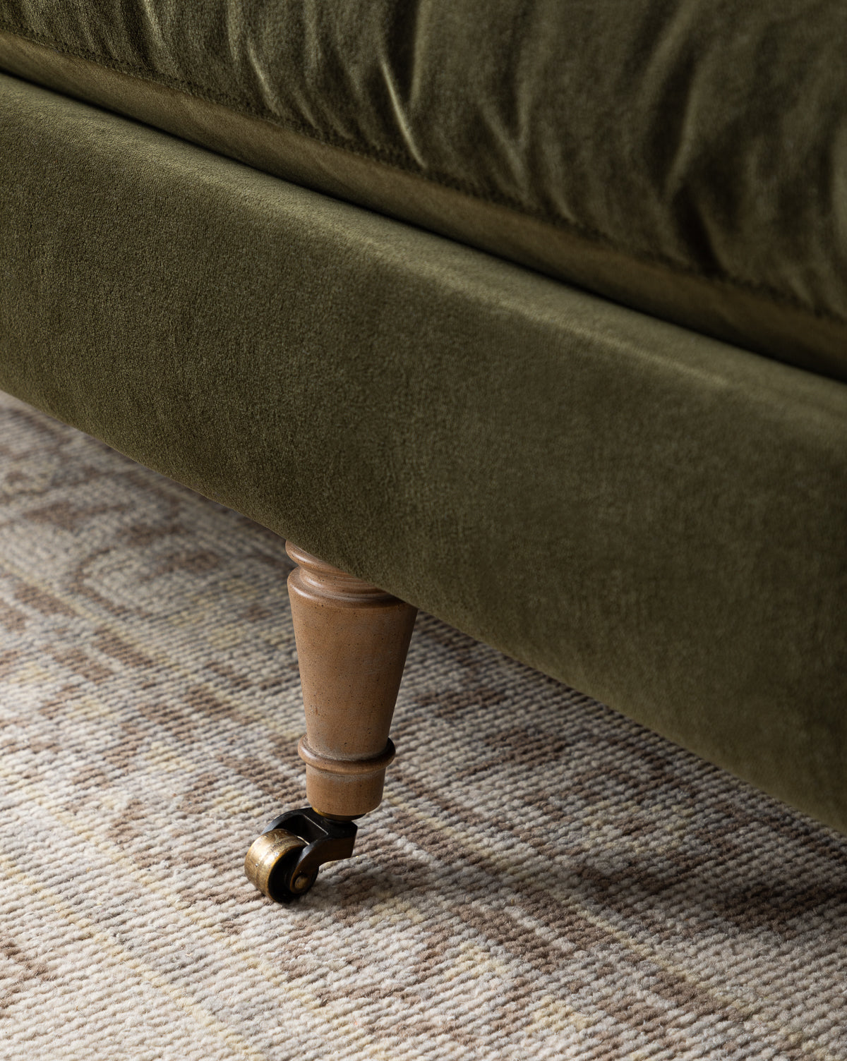 Close-up of a handcrafted sofa leg with a brass caster wheel, attached to the Haverford Upholstered Sofa 90 in olive green, resting on a beige and brown patterned rug by McGee & Co.