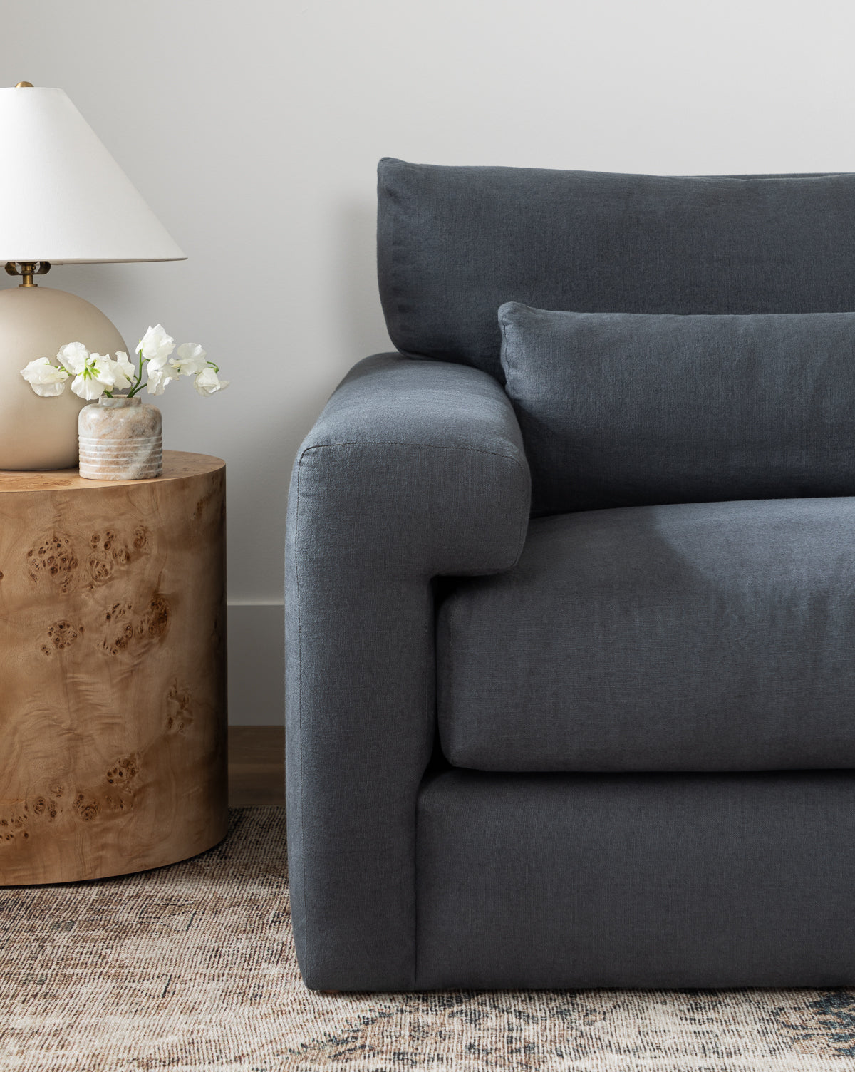 A modern living room featuring the Community Beckstead Chaise Sectional in dark gray, paired with a round wooden side table, a beige lamp, and a small vase of white flowers atop a neutral patterned rug, finished with touches from McGee & Co.