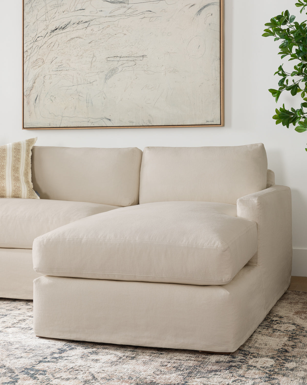 A modern living room features the Community Macy Slipcover Chaise Sectional 101 in beige, paired with a patterned throw pillow, an abstract wall painting, a green plant, and a neutral-toned area rug from McGee & Co.