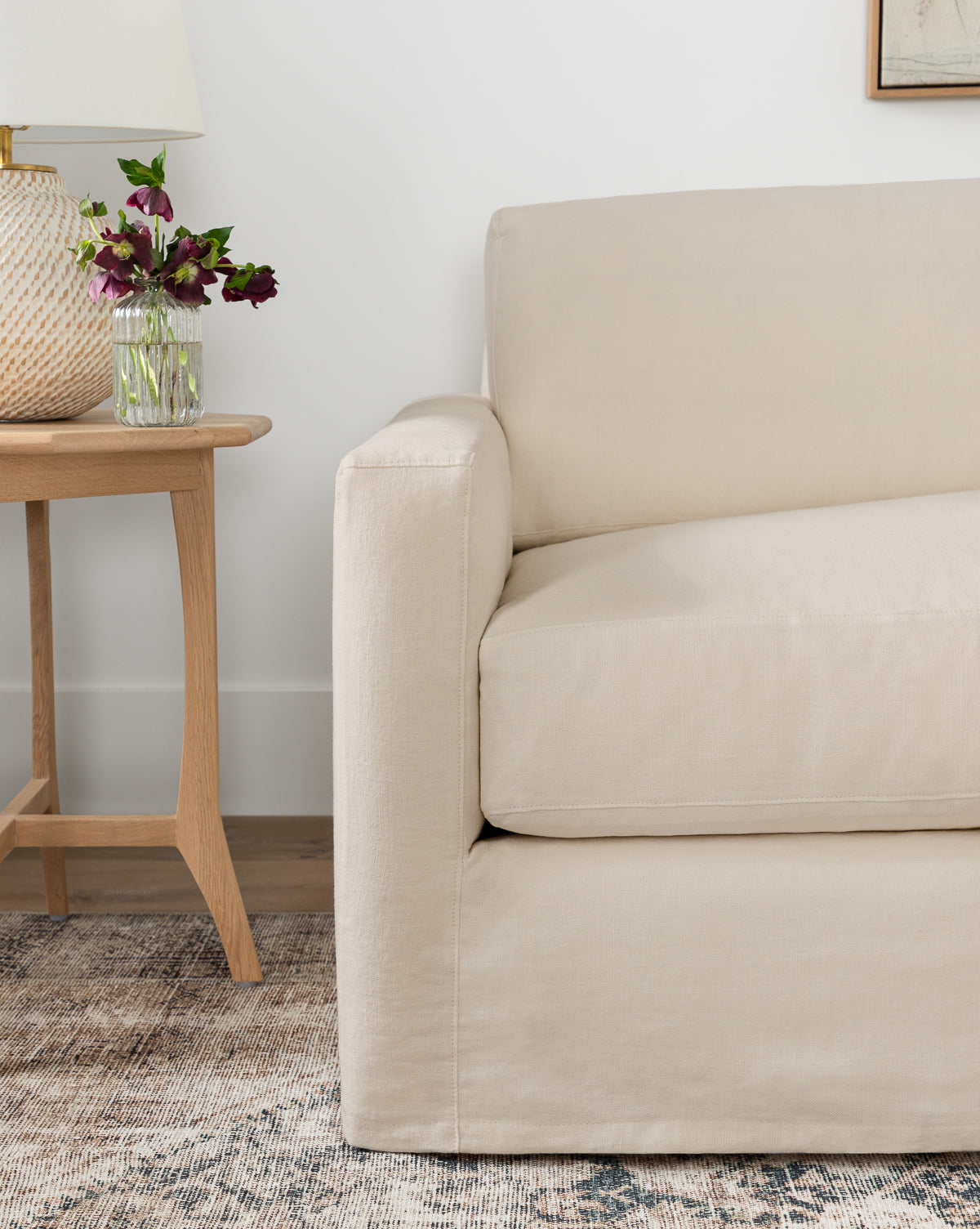 A beige upholstered armchair sits by a wooden side table with a glass vase of purple flowers and a white lamp, on a neutral patterned rug—perfectly complementing the Community Macy Slipcover Chaise Sectional 101 by McGee & Co.