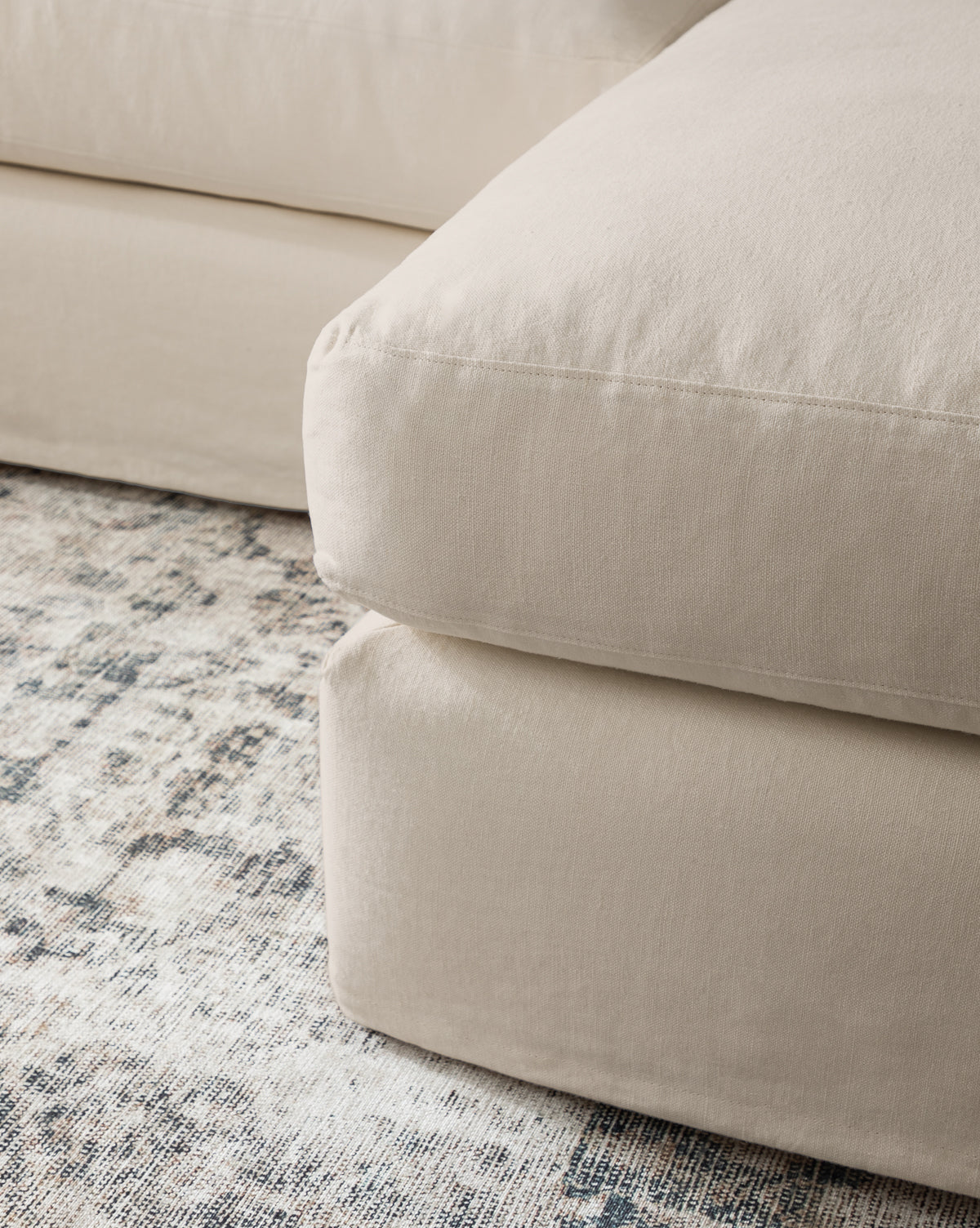 A close-up of the Community Macy Slipcover Chaise Sectional 101 in beige upholstery with large, plush cushions, set on a textured, patterned neutral-toned area rug, McGee & Co.