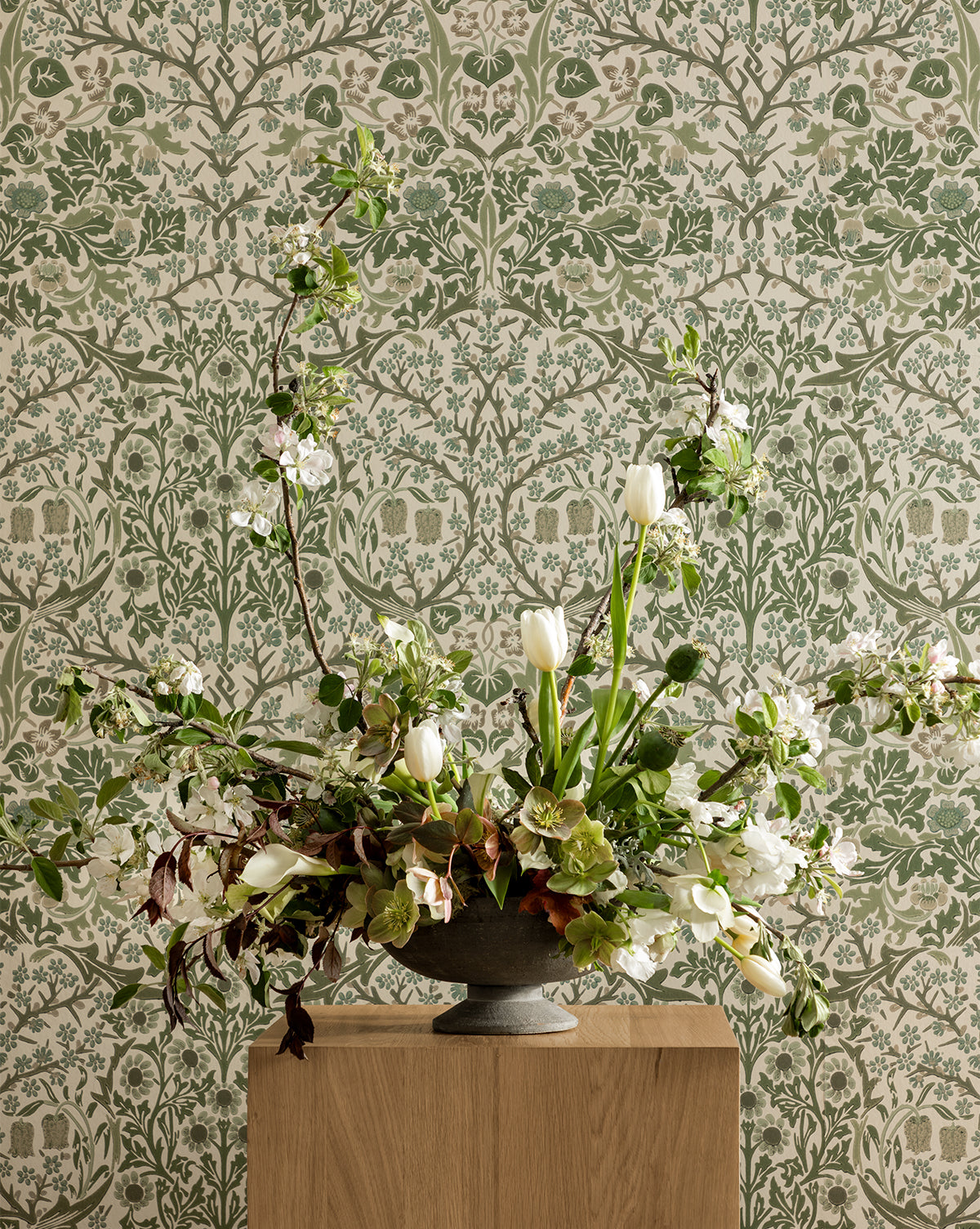 A large floral arrangement with white tulips and greenery in a gray vase sits on a wooden pedestal, set against Morris & Co. x McGee & Co. Blackthorn Wallpaper by Sanderson Design Group, Inc., featuring a botanical green and cream design by McGee & Co.