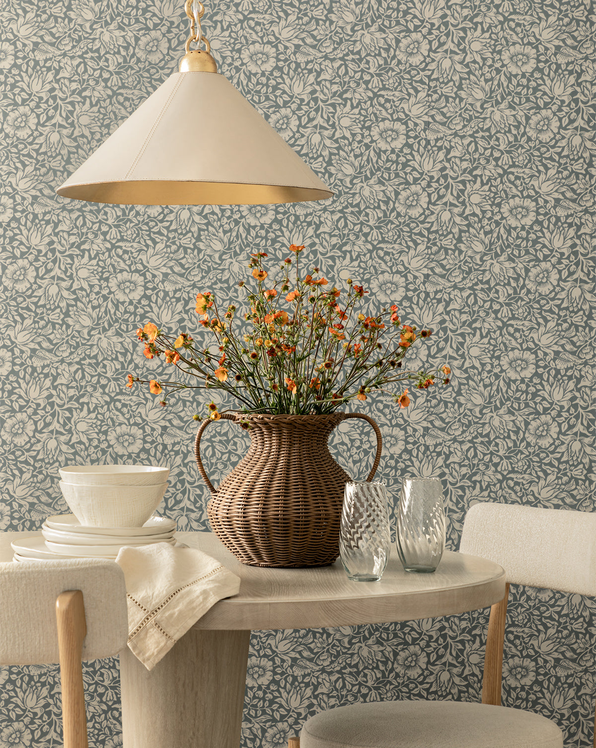 A round table with a beige tablecloth, two white chairs, the Antonia Woven Vase filled with orange flowers, two textured glasses, stacked white bowls, and a pendant light set against floral-patterned wallpaper.