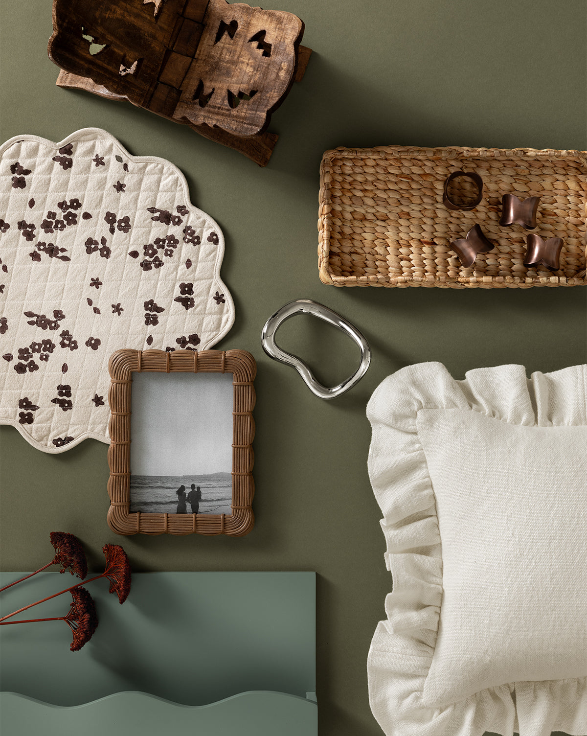 A flat lay of home decor on a green surface, including a ruffled cream pillow, wicker basket, wooden trinket box, metal object, dried flowers, floral placemat, green wooden tray with organic texture, and the Hilde Woven Frame.