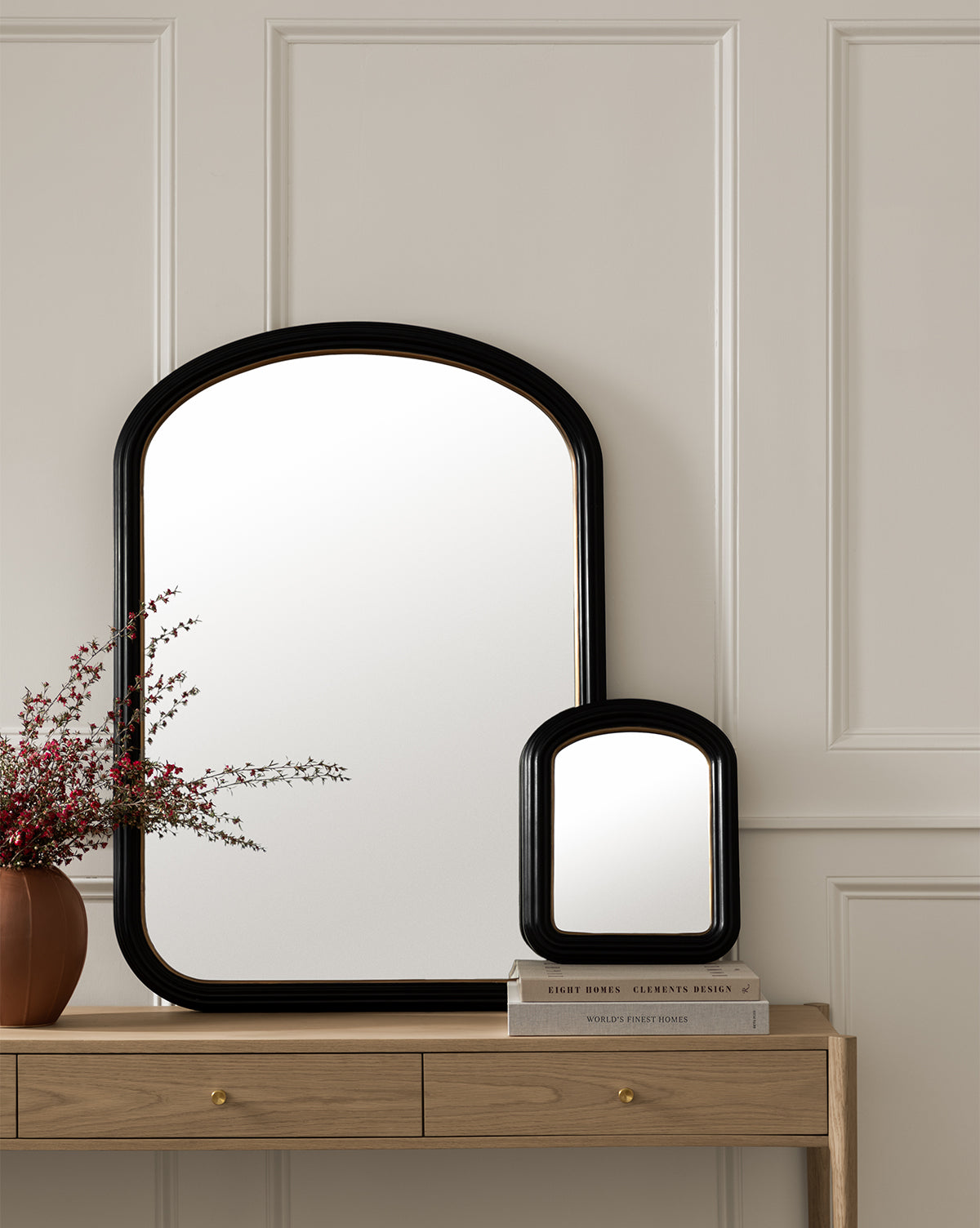Two Samuel Shelf Mirrors with black finish rest on a light wood console table against a white paneled wall. A brown vase with red and white flowers and stacked books complete the arrangement, by McGee & Co.