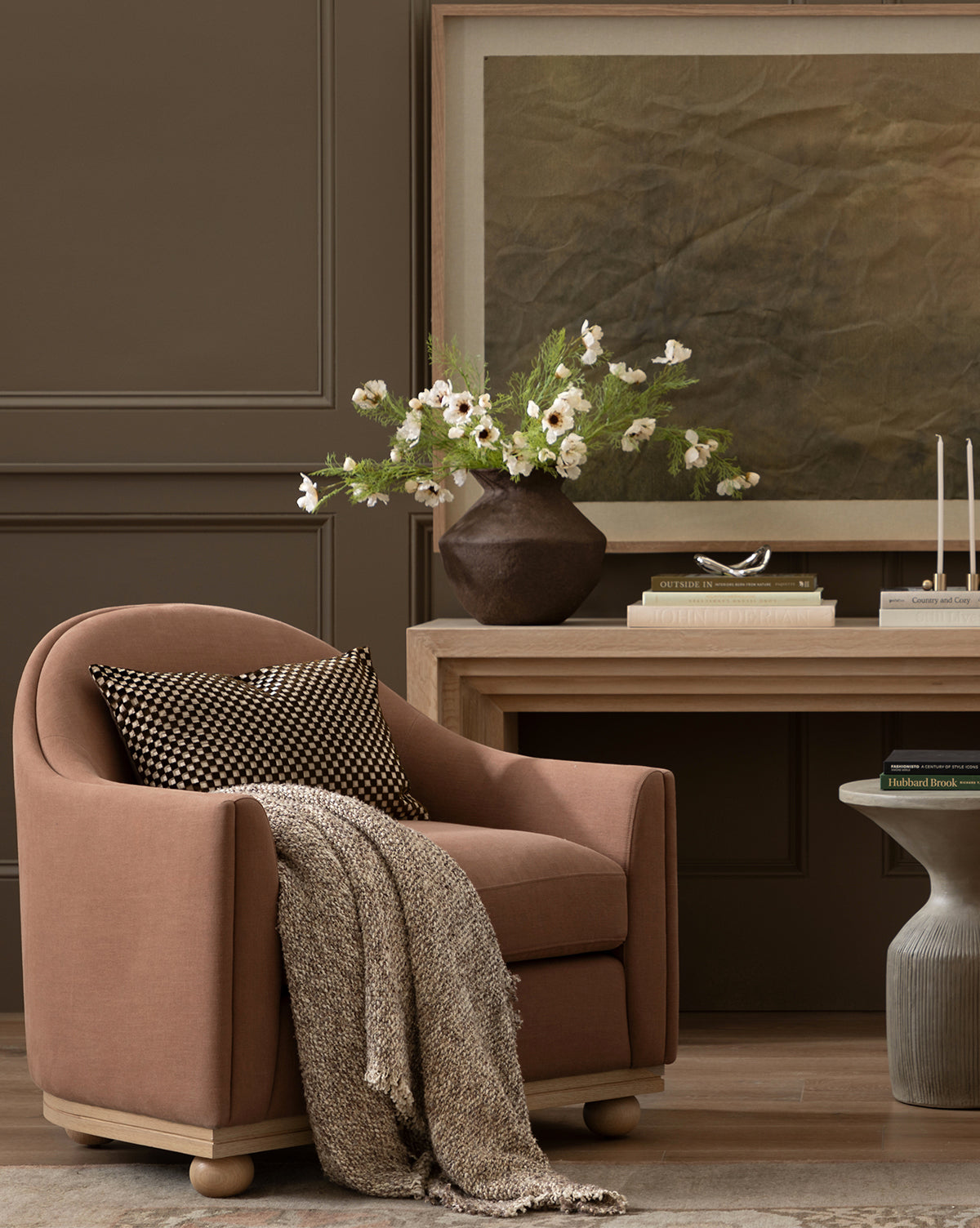 A mauve armchair with a throw and cushion sits beside a wooden desk. On the desk is a large vase of Cream Cosmos Stem, candles, and abstract art, all set against a brown paneled wall by McGee & Co.