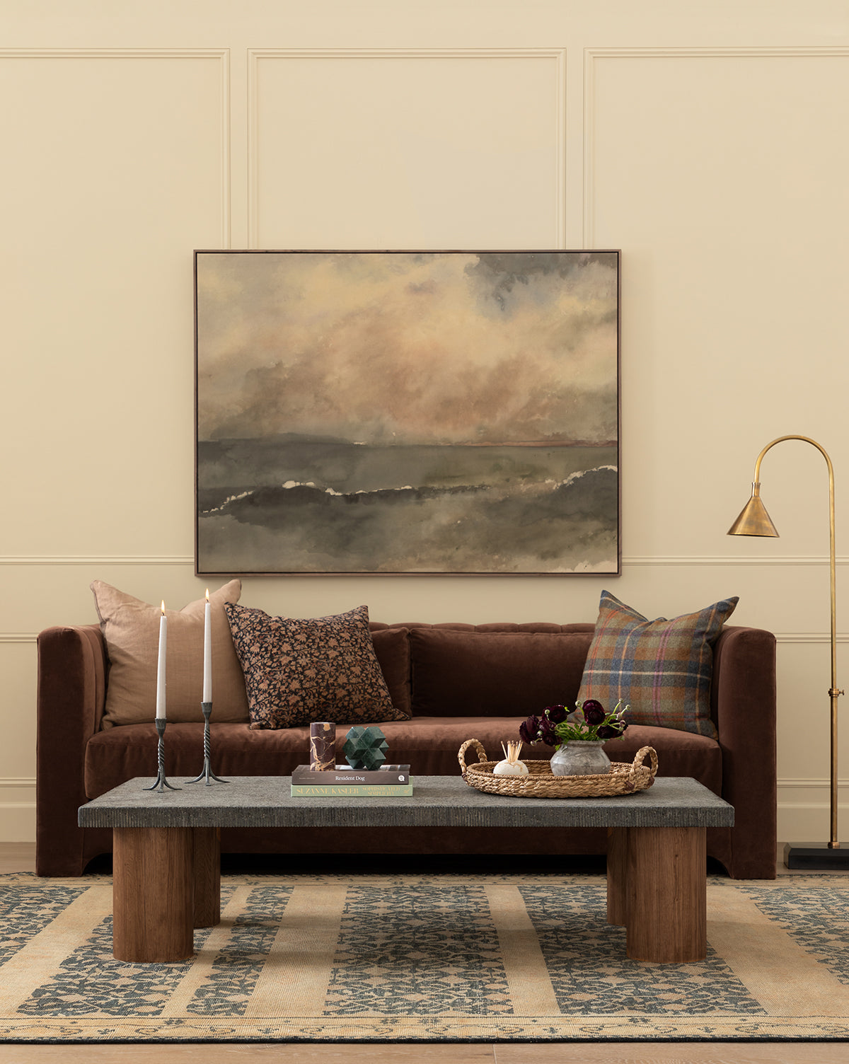 A cozy living room features a brown sofa with patterned pillows, a wooden coffee table with decor, tall candles, the gallery-quality You Said I am the Sea by Aileen Fitzgerald painting, a brass floor lamp, and a blue patterned rug on beige flooring, by McGee & Co.