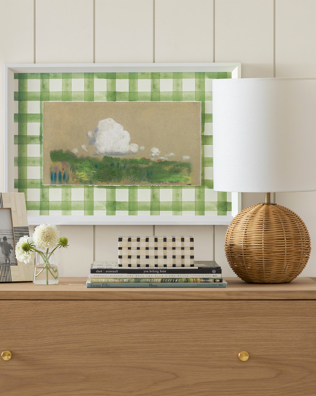 A wooden dresser displays Home: a wicker table lamp with a white shade, stacked books including a Carson Ellis children’s book, a small glass vase, and a black-and-white framed photo. Above hangs a green gingham-framed landscape painting on the white paneled wall.