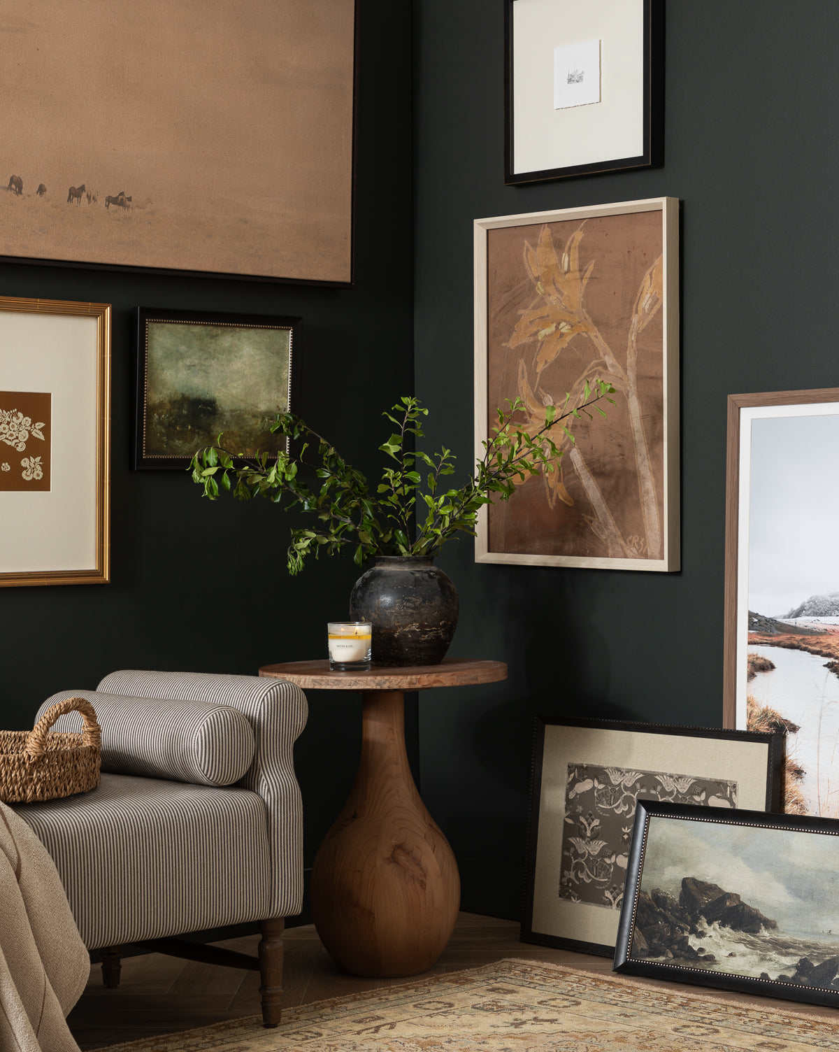 A cozy living space with dark walls features unique wall treatments, a round wooden side table with a black vase of green branches and a candle, and framed art—including the Blowing Brush gallery-wrapped canvas—displayed on floor and wall, by McGee & Co.