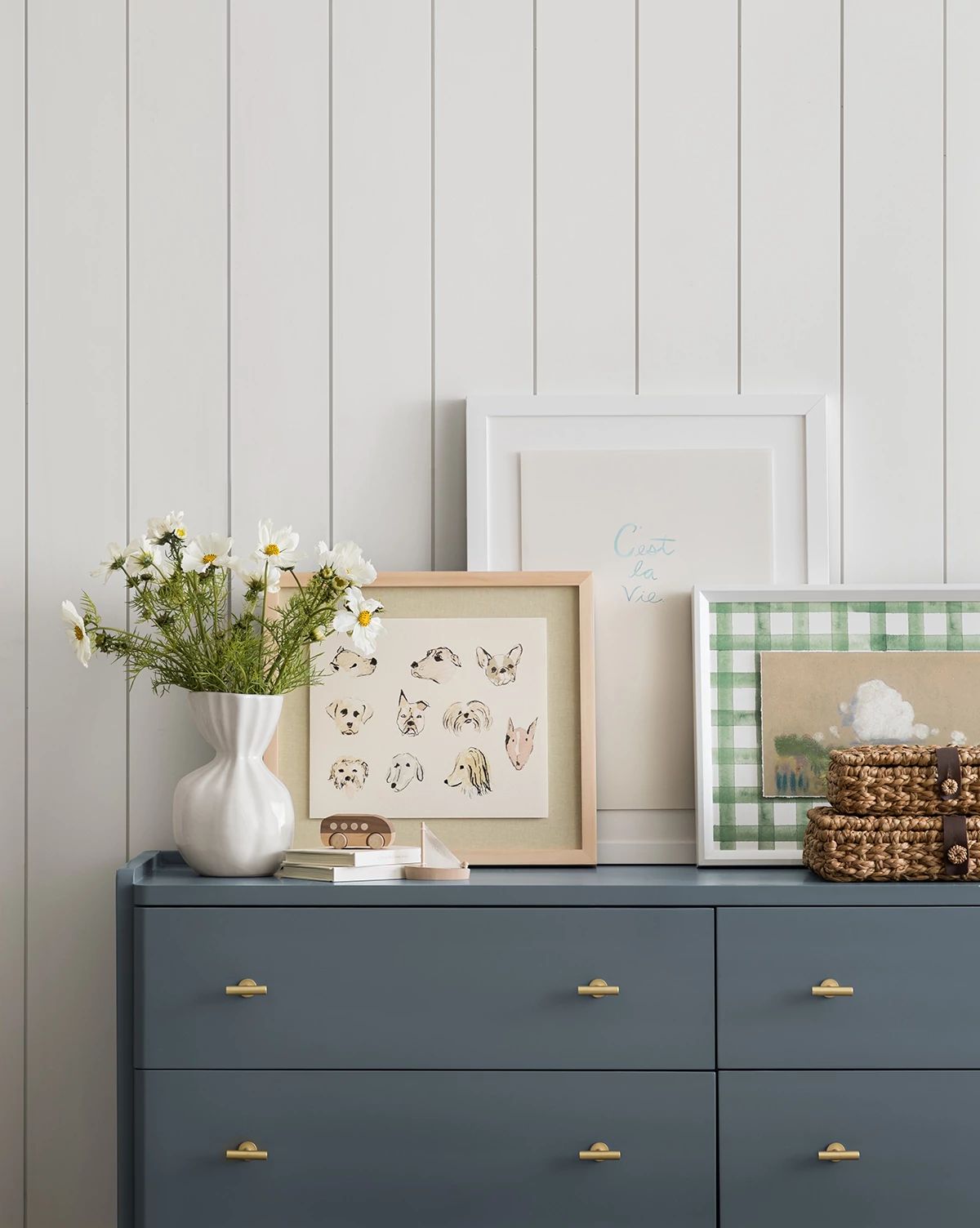 A blue dresser with gold handles displays a white vase of daisies, framed art, woven baskets, and the Woof ceramic dog dish against a white shiplap wall, by McGee & Co.