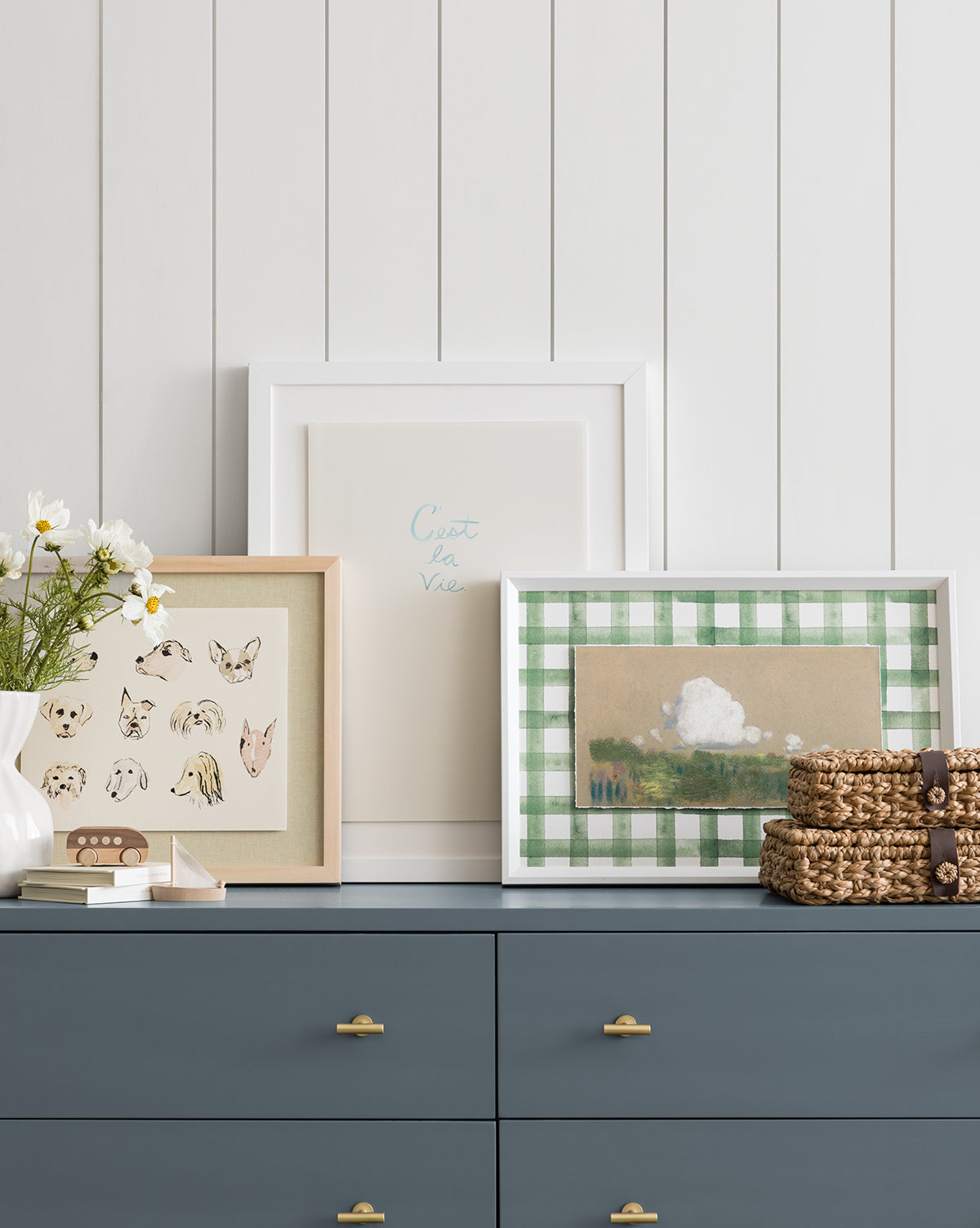 Dreamscape, a blue dresser with gold handles, displays a vase of daisies, a dish, woven coasters, and three framed artworks—featuring sketches, text, and a checkered landscape—set against a white paneled wall for stylish home decor, by McGee & Co.