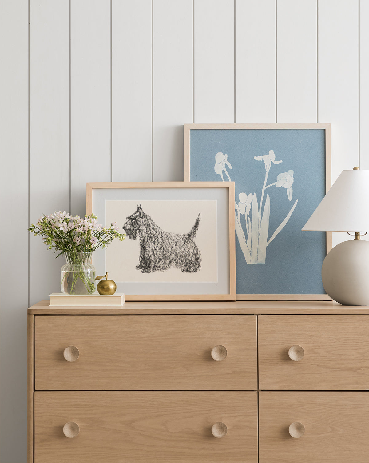 The Finn light wood dresser with round knobs holds a small glass vase of flowers, a gold trinket, and a white table lamp. Two framed artworks—a Scottish Terrier sketch and white blooms on blue—lean against a white paneled wall, by McGee & Co.