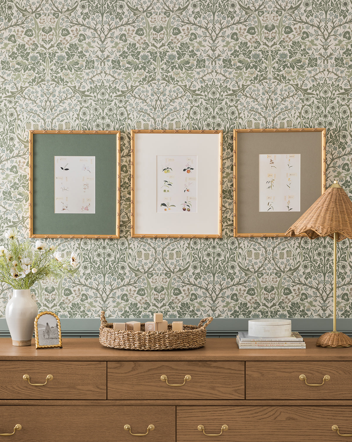 A wooden dresser with gold handles stands before floral wallpaper. Above it hang three Botanical Grid 3 gallery-quality prints. On the dresser are a flower vase, framed photo, woven basket, and a lamp with a pleated shade, by McGee & Co.