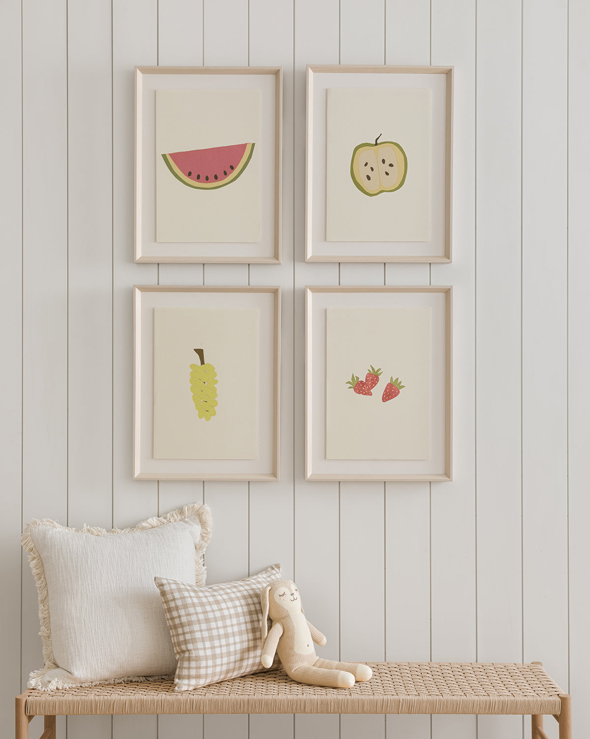 A cozy bench with two pillows and a plush bunny sits against a white shiplap wall. Above, four framed art prints—including the Apple Collage giclee print and other fruit-themed illustrations—are arranged in a grid by McGee & Co.
