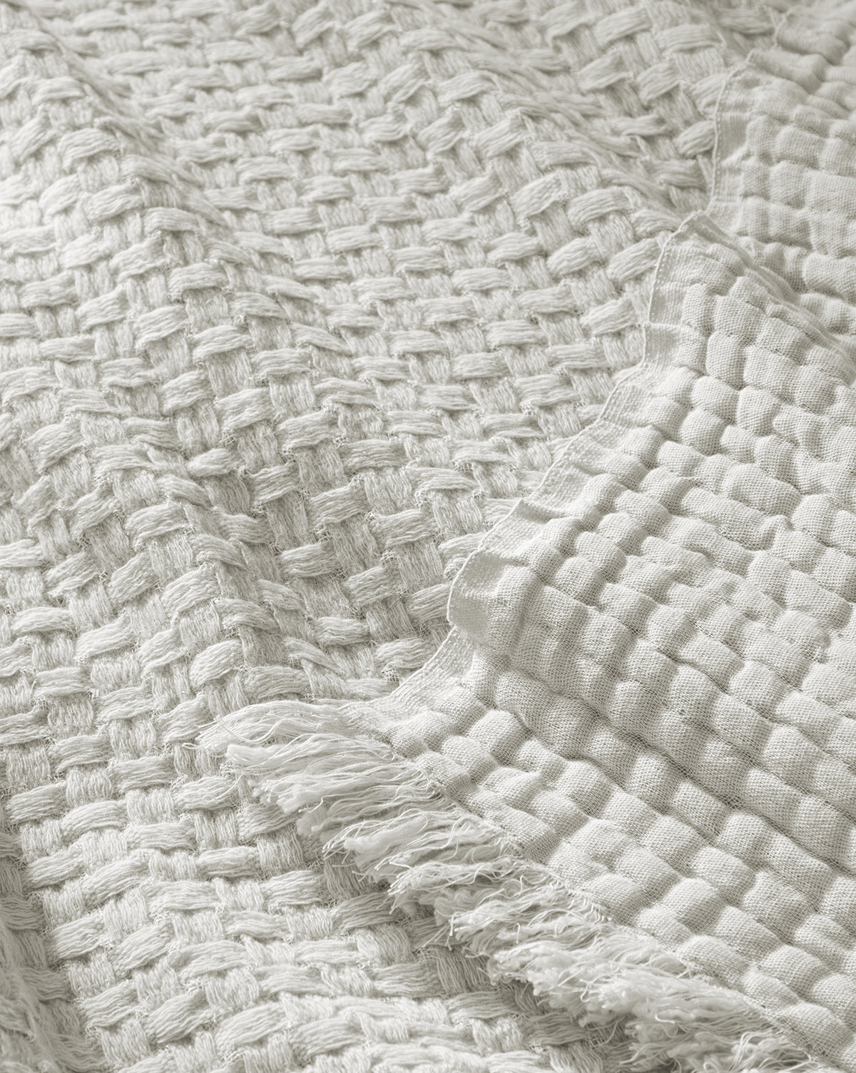 A close-up of two off-white Caroline Kids Blankets by Indigo Craft—one shows a thick basket weave, the other a finer grid pattern with frayed edges, both resembling soft woven textures. McGee & Co.