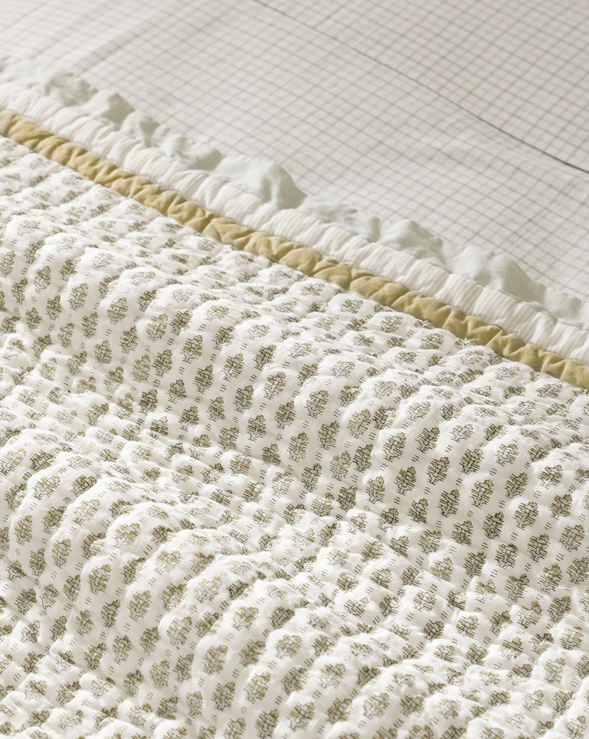 A close-up of the Dulcie Block Print Kids Quilt by Renuka Inc., showcasing a delicate leaf pattern in light brown on cream, ruffled and textured details, with a grid-patterned sheet beneath—crafted in soft cotton quilt style by McGee & Co.