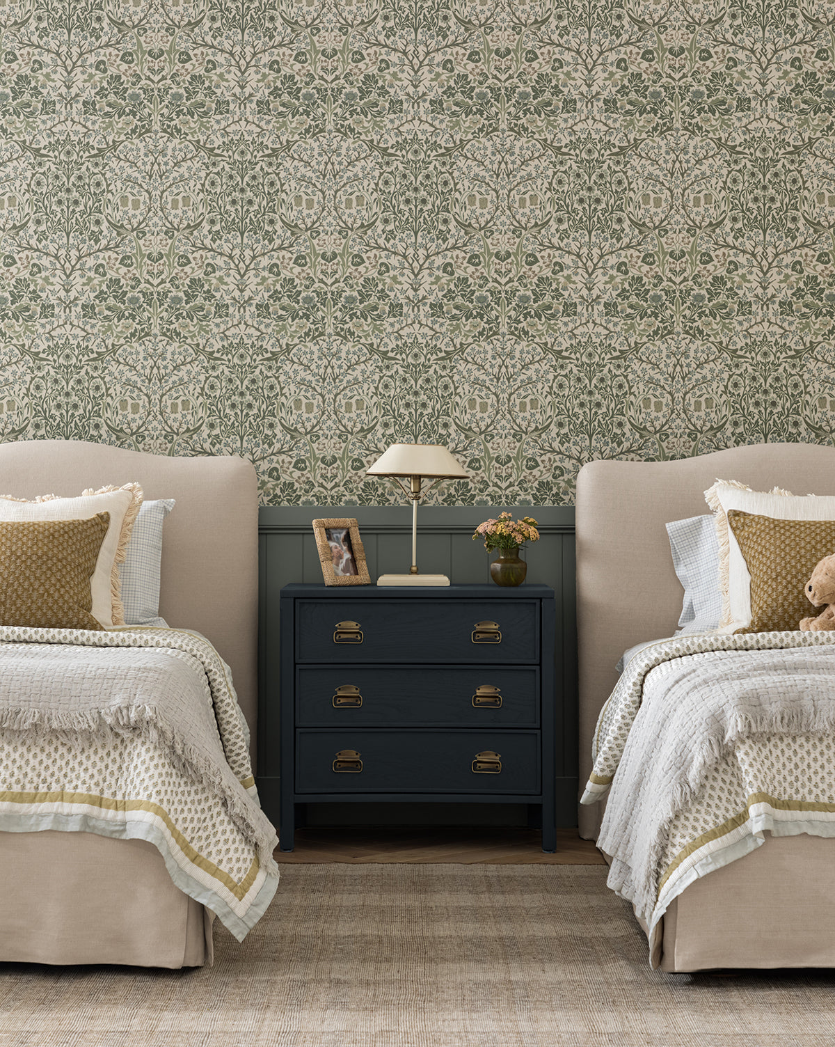 Two twin beds with beige headboards and bedding are separated by a navy nightstand with a lamp, photo, and book. Behind them, Sanderson Design Group’s Morris & Co. x McGee & Co. Blackthorn Wallpaper adds green floral charm; a teddy bear sits on one bed—a cozy space layered with thoughtful details from McGee & Co.