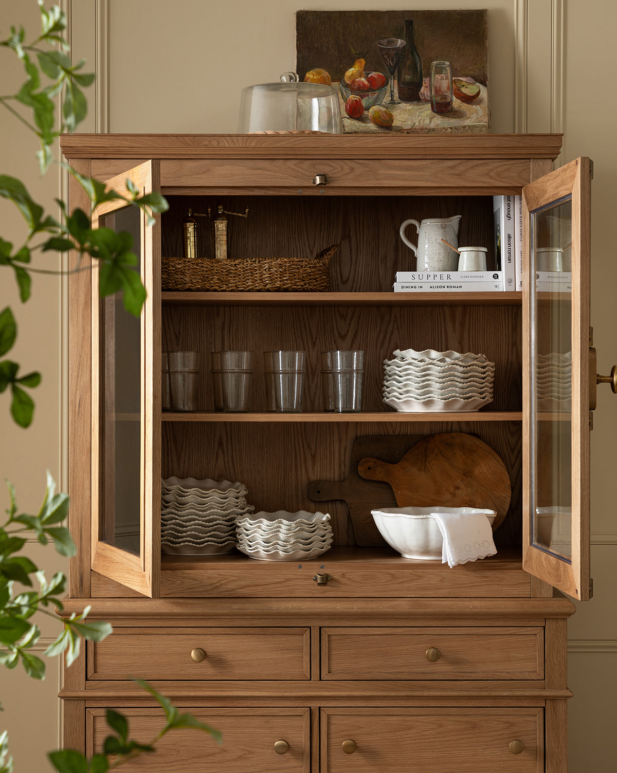The Heath Oak Cabinet with glass doors displays wavy white dishes, clear glasses, books, a pitcher, bowl, and baskets. On top rest a cutting board and fruit still-life painting; greenery is visible at the side, by McGee & Co.