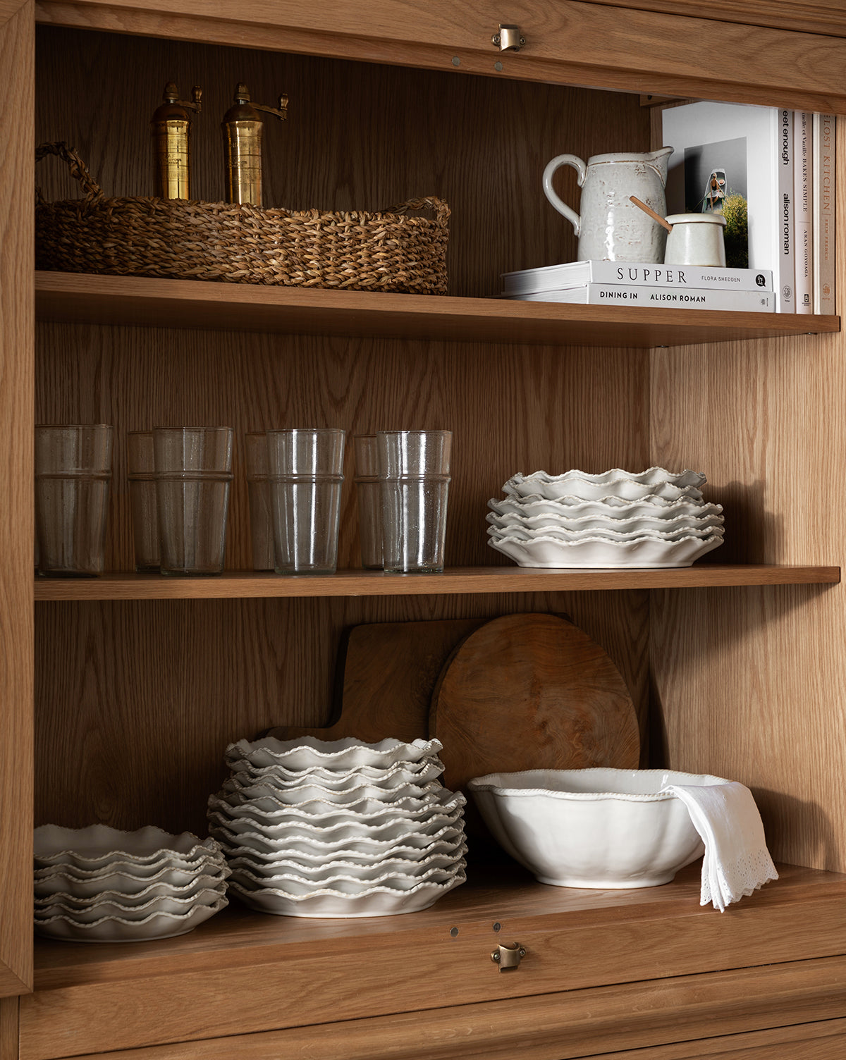 A wooden cabinet with shelves holding glass tumblers, wavy-edged white plates, bowls including the McGee & Co. x Loeffler Randall Tabitha Serving Bowl, wooden cutting boards, a basket with bottles, a white pitcher, and neatly stacked books by McGee & Co.