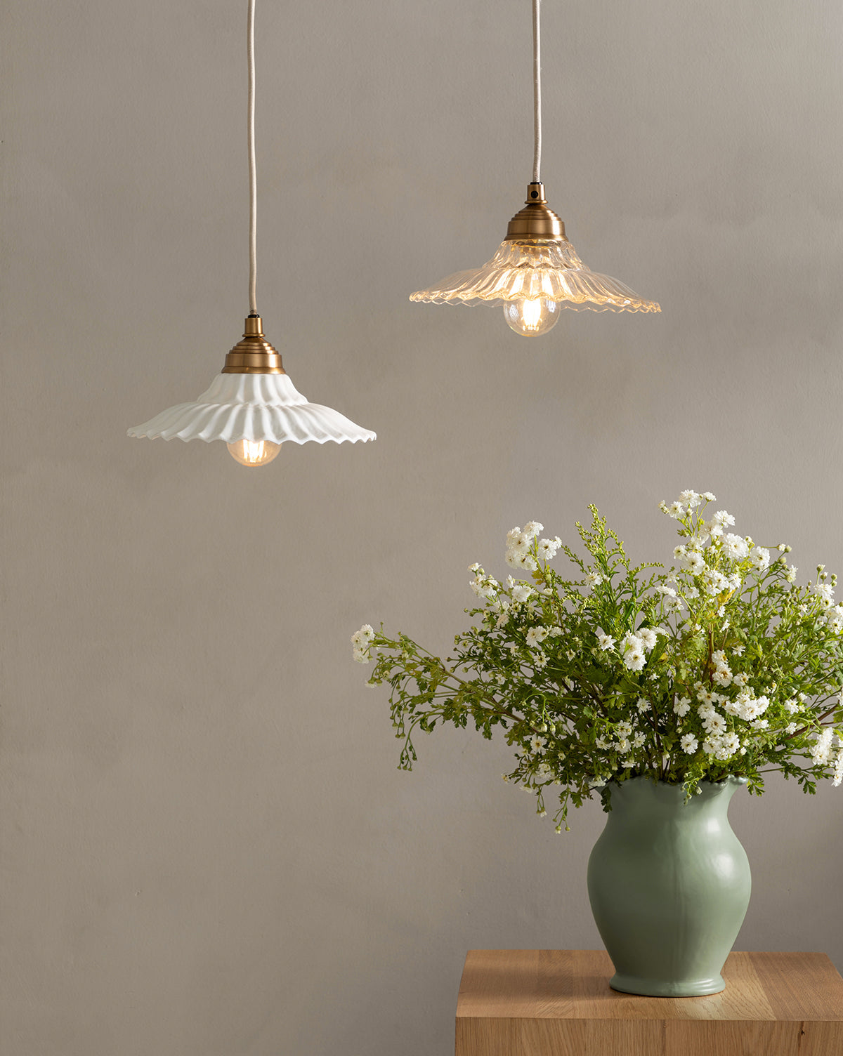 Two McGee & Co. x Loeffler Randall Maude Glass Pendant lights with scalloped glass shades hang above a green vase filled with white flowers and greenery on a wooden table against a plain light gray wall, by McGee & Co.
