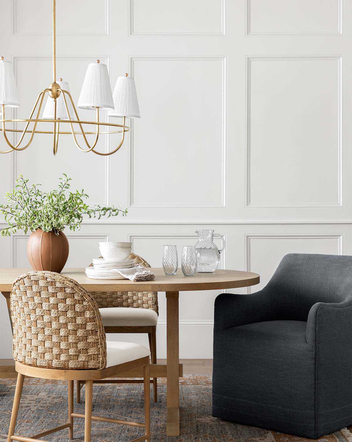 A modern dining area showcases a round wooden table with dishes, glassware, and a pitcher. The space features a woven chair, an Elton Dining Chair, a potted plant, and a gold chandelier set against white paneled walls, by McGee & Co.