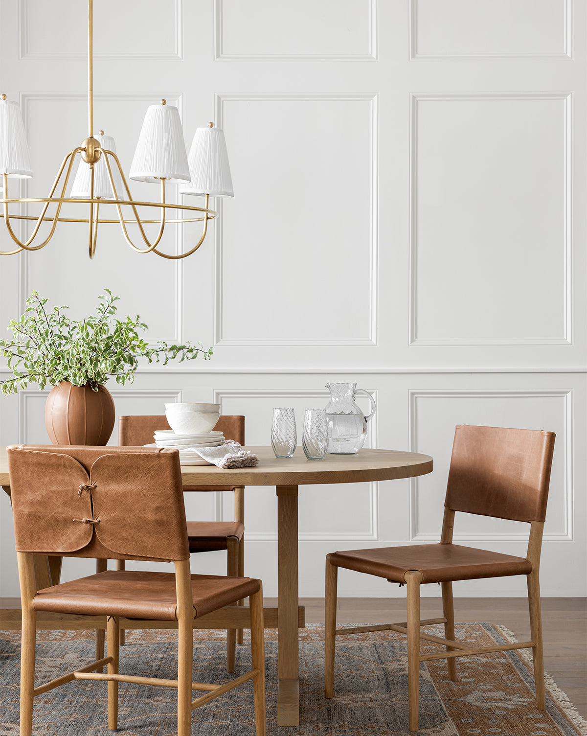 A modern dining area features a round wooden table, four Danson Dining Chairs with brown leather seats, tableware, a potted plant, and a gold chandelier against a white paneled wall for a minimalist look, by McGee & Co.